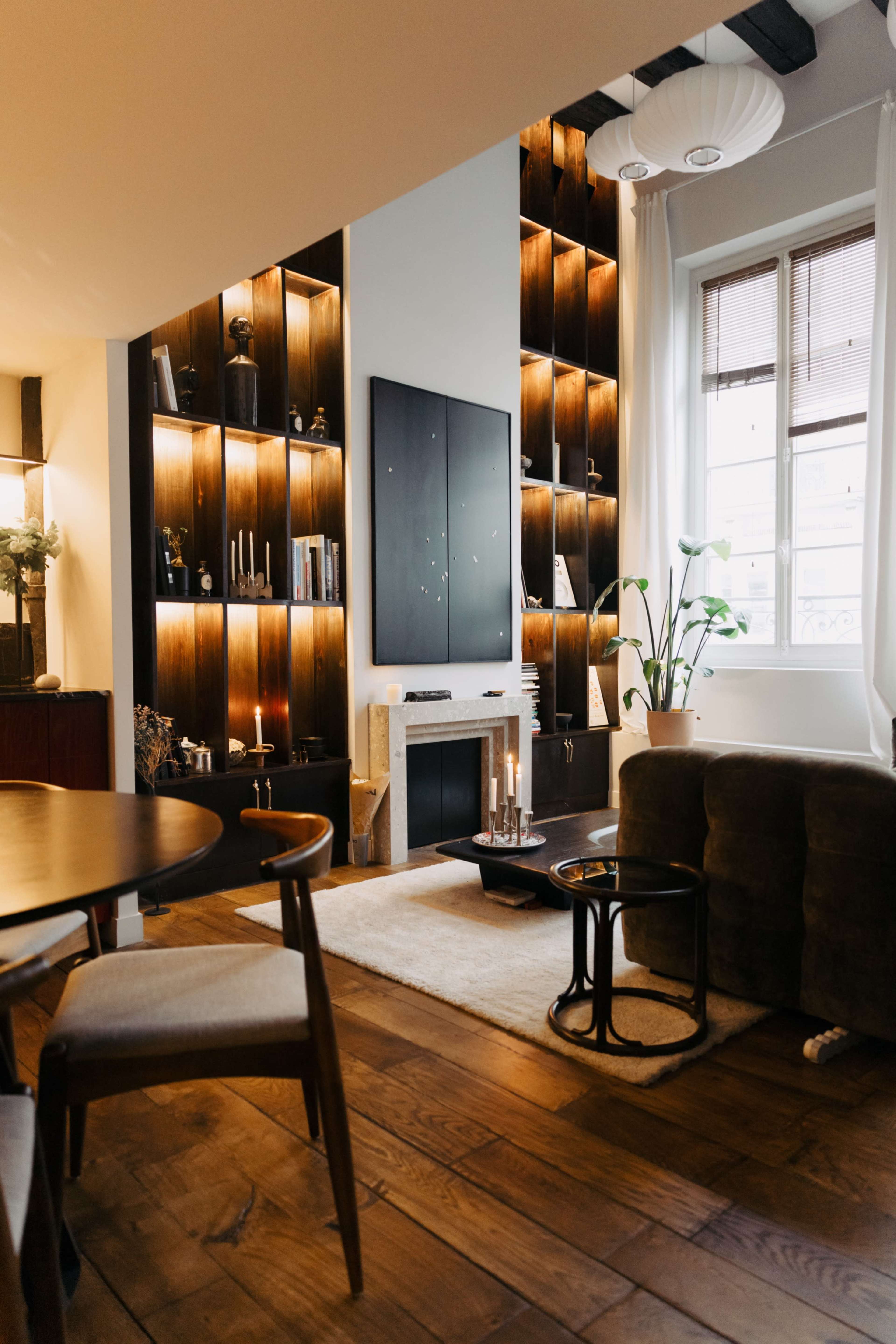 The image shows a cozy living room featuring dark wood shelves with decorative items, a fireplace, a plush sofa, and a window letting in natural light.