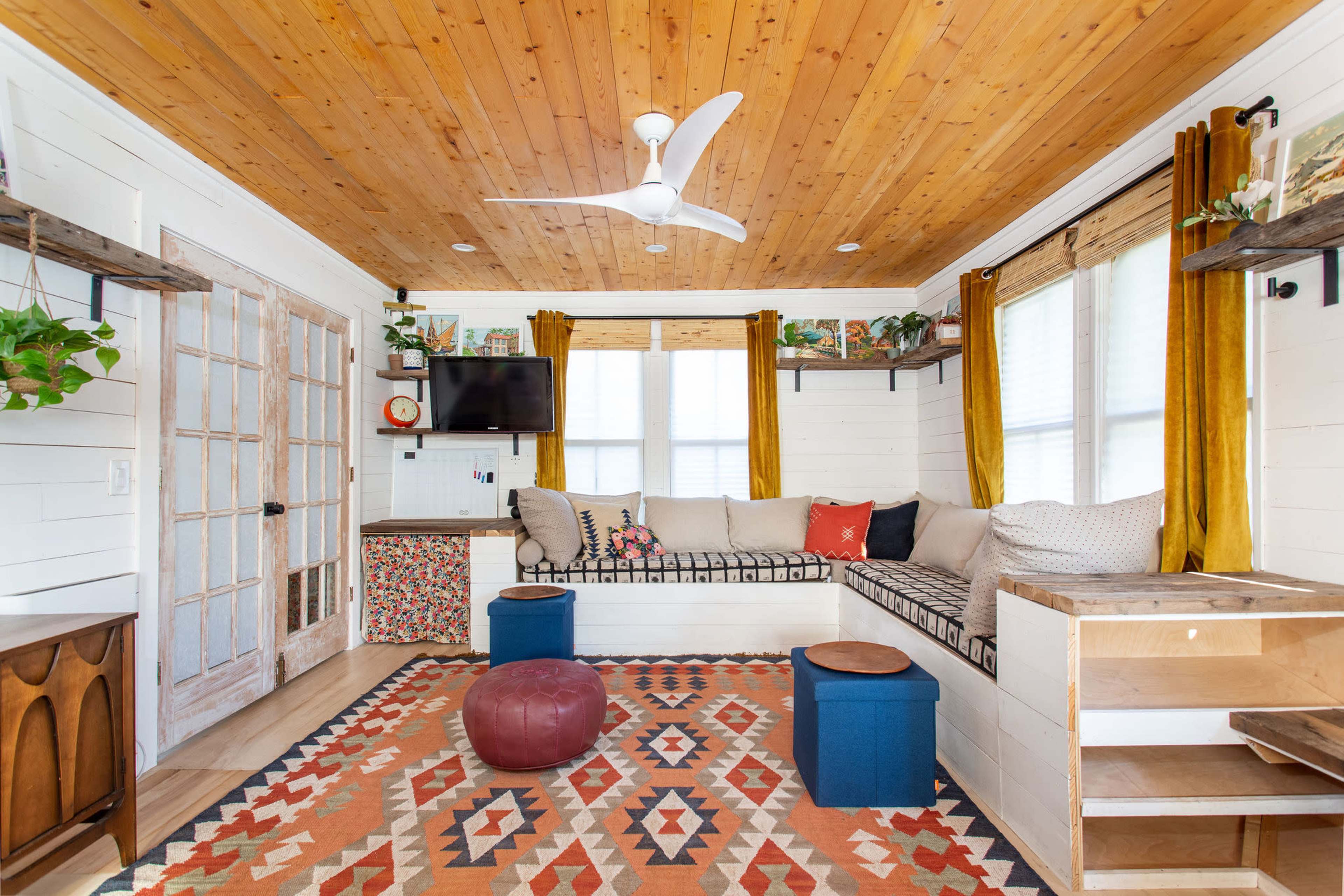 A cozy living room with a wooden ceiling, a sectional sofa, a patterned rug, and decorative shelves displaying plants and items.
