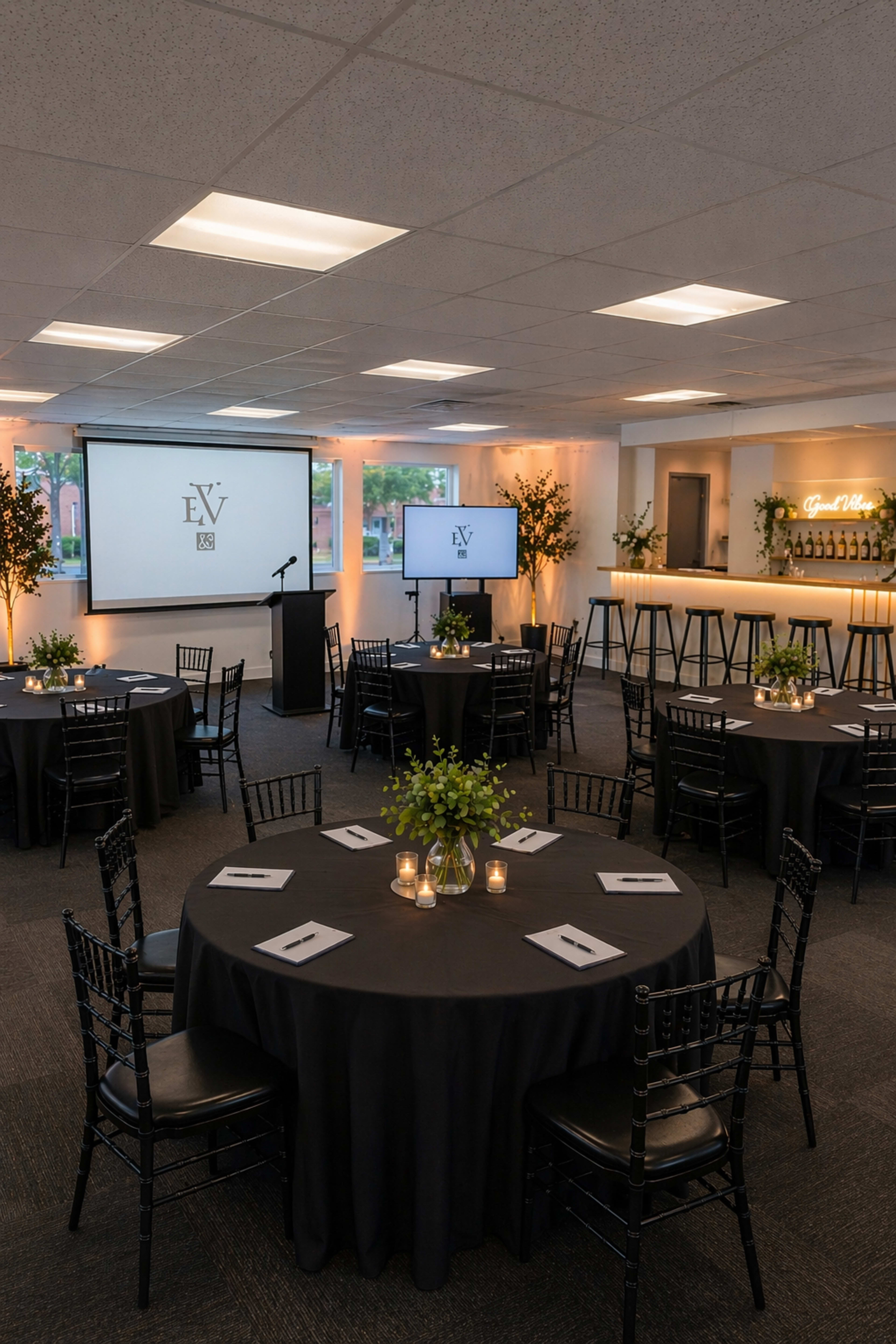 The image shows an event space set up with round tables covered in black tablecloths, each featuring centerpieces, candles, and a podium with a presentation screen in the background.
