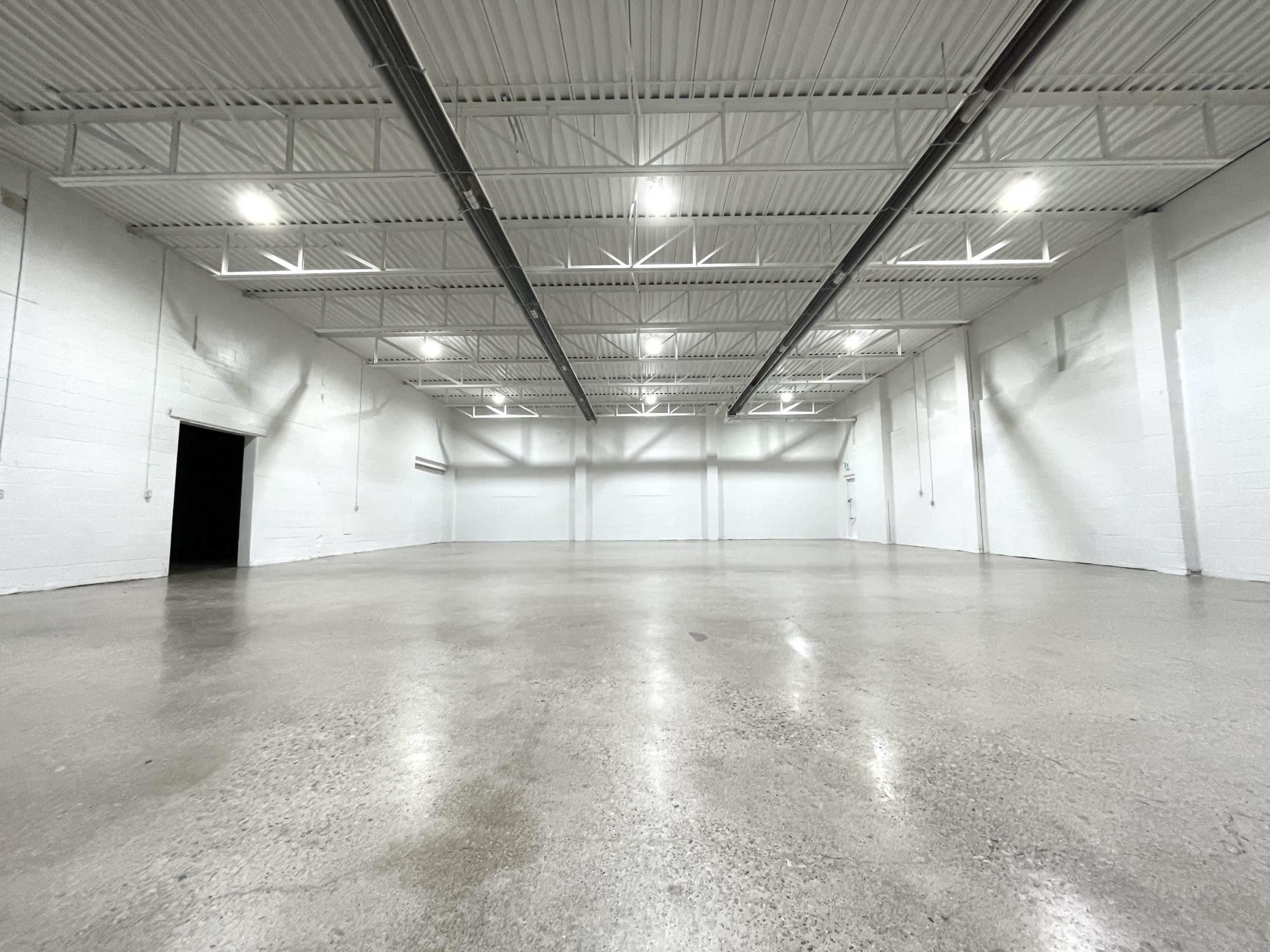 The image shows a large, empty warehouse with a polished concrete floor and white walls, illuminated by overhead lights.
