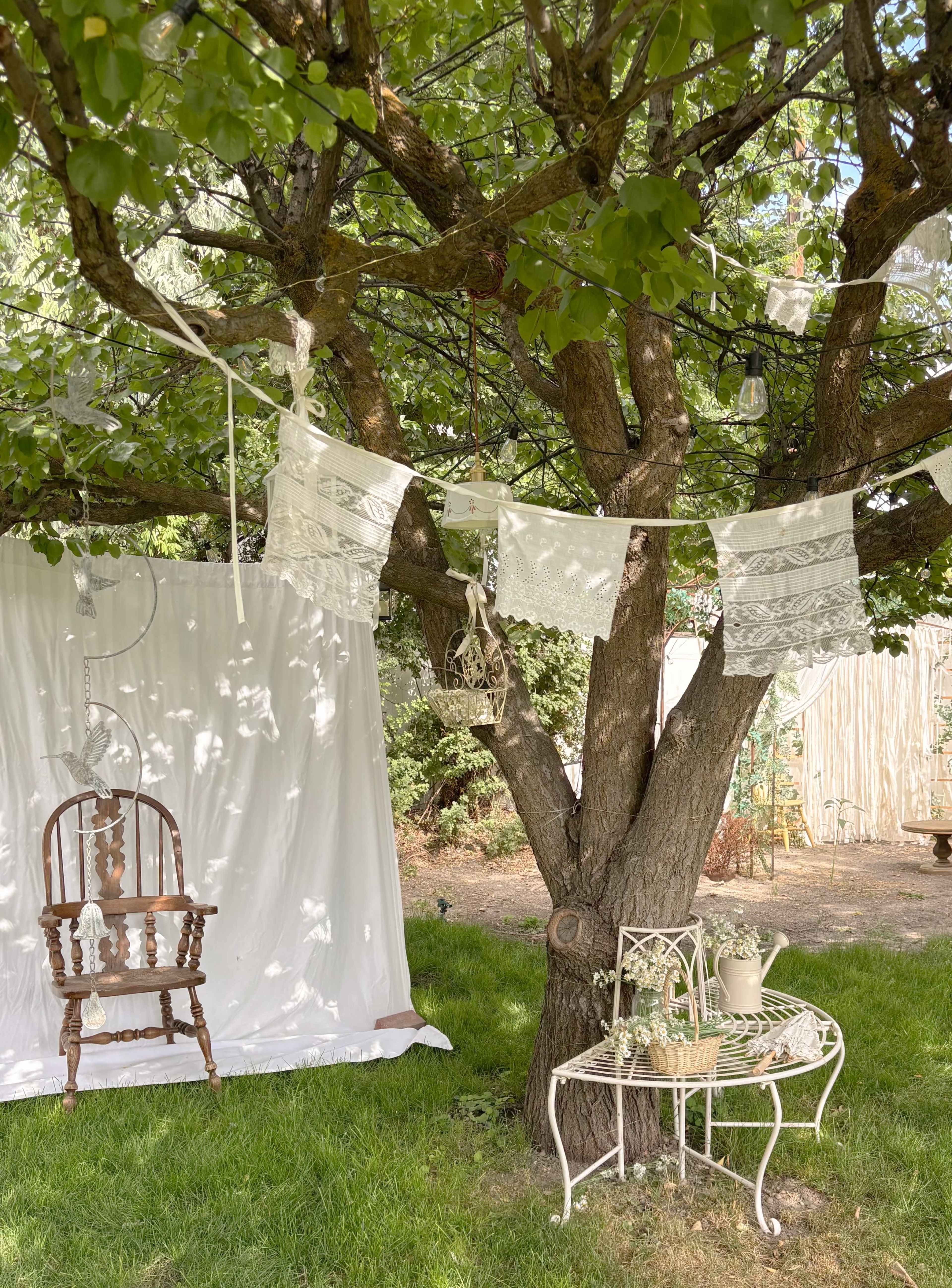 A rustic outdoor scene featuring a large tree with white fabric draped nearby, adorned with decorative banners and a vintage chair set against a grassy backdrop.
