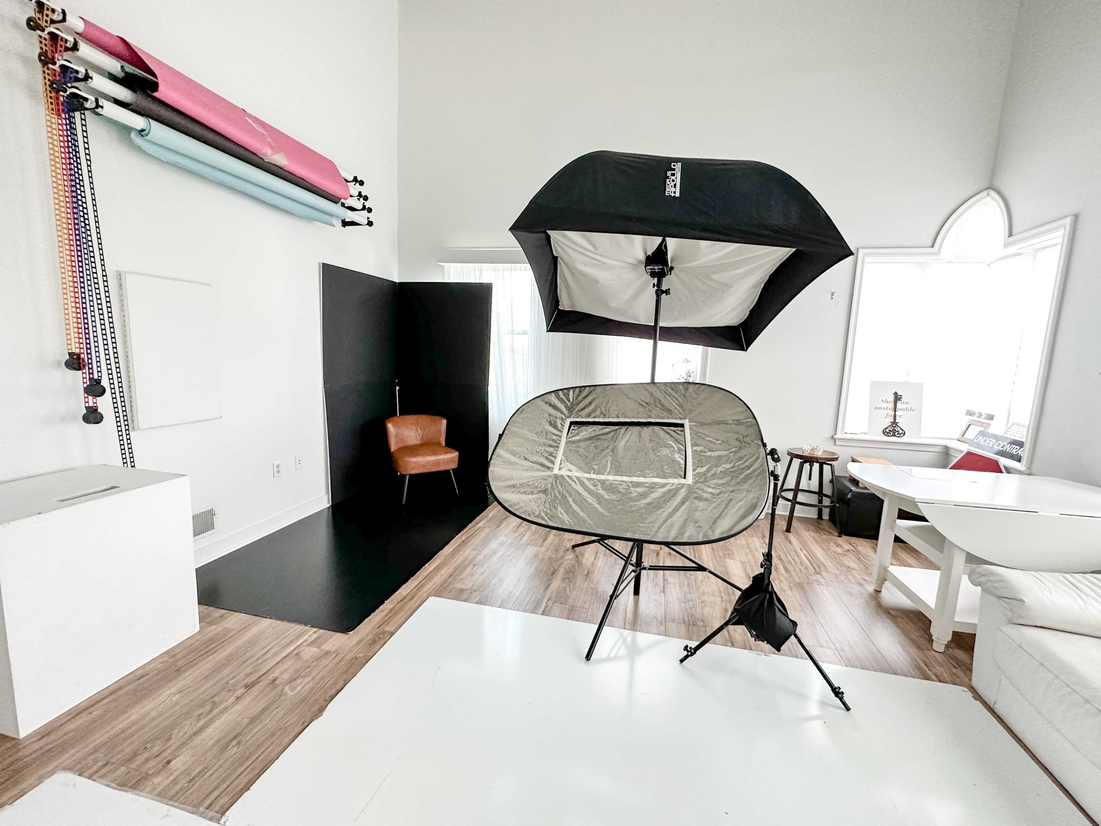 A photography studio setup featuring a light umbrella, a portable backdrop, a wooden chair, and various backdrops on the wall.