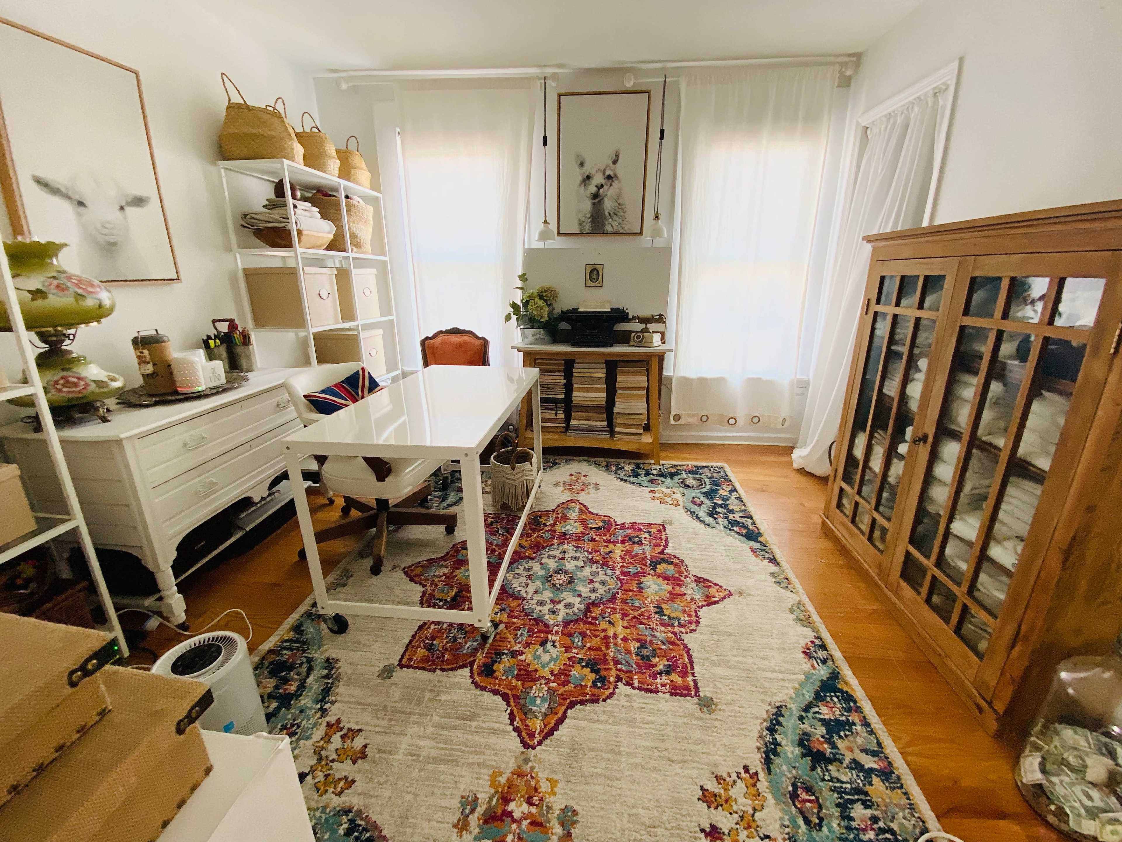 A bright room with a white desk, bookshelf, and a wooden cabinet, all arranged on a colorful patterned rug.
