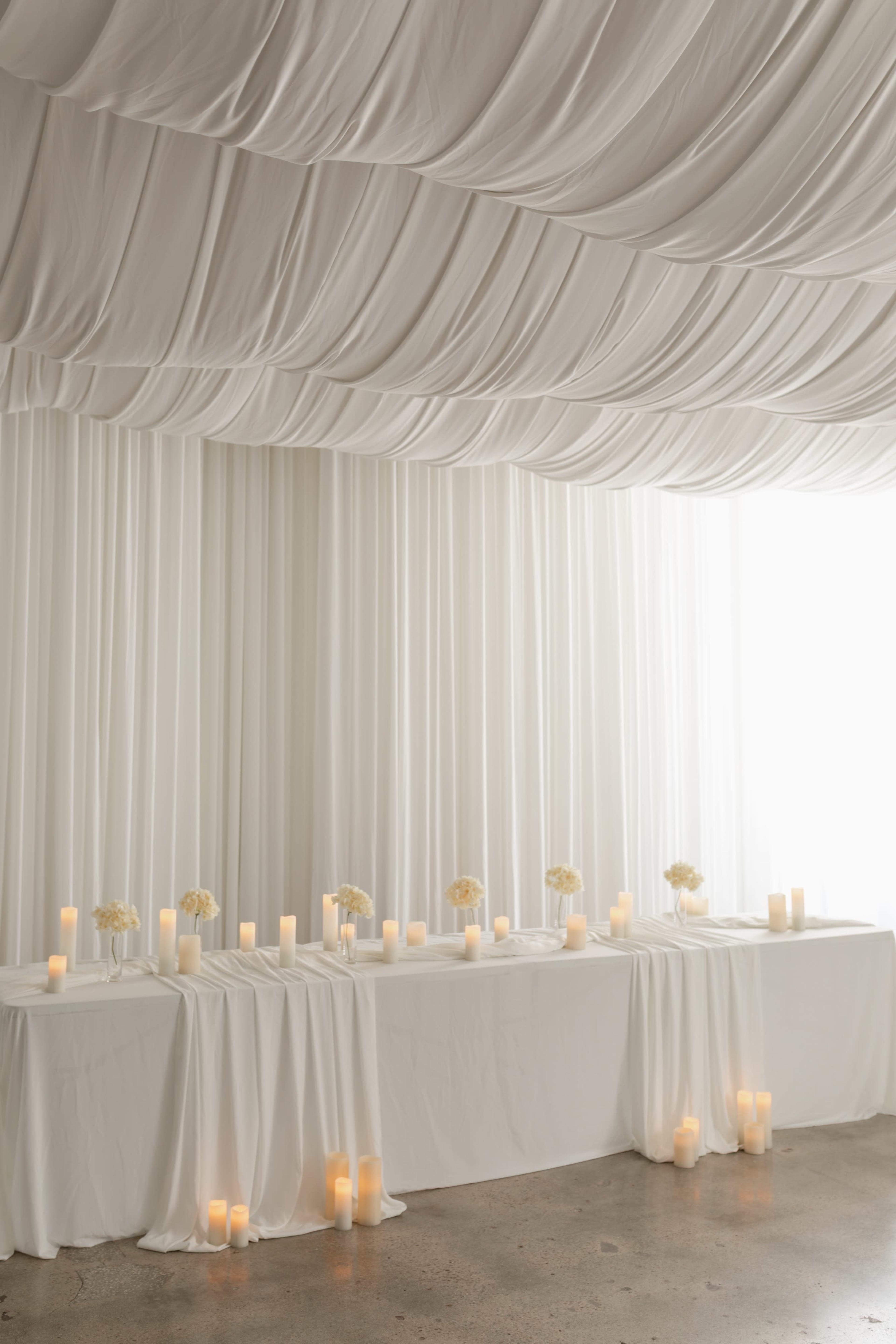 The image shows a softly lit, elegantly draped white backdrop with a long table adorned with candles and small floral arrangements.