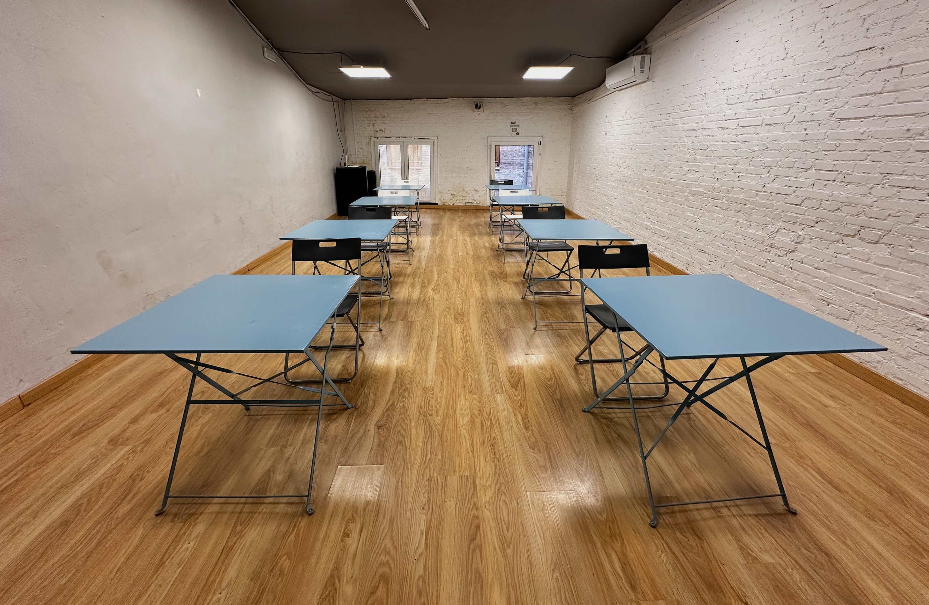 A neatly arranged classroom features six blue folding tables spaced evenly on a wooden floor, with a blank white wall in the background.