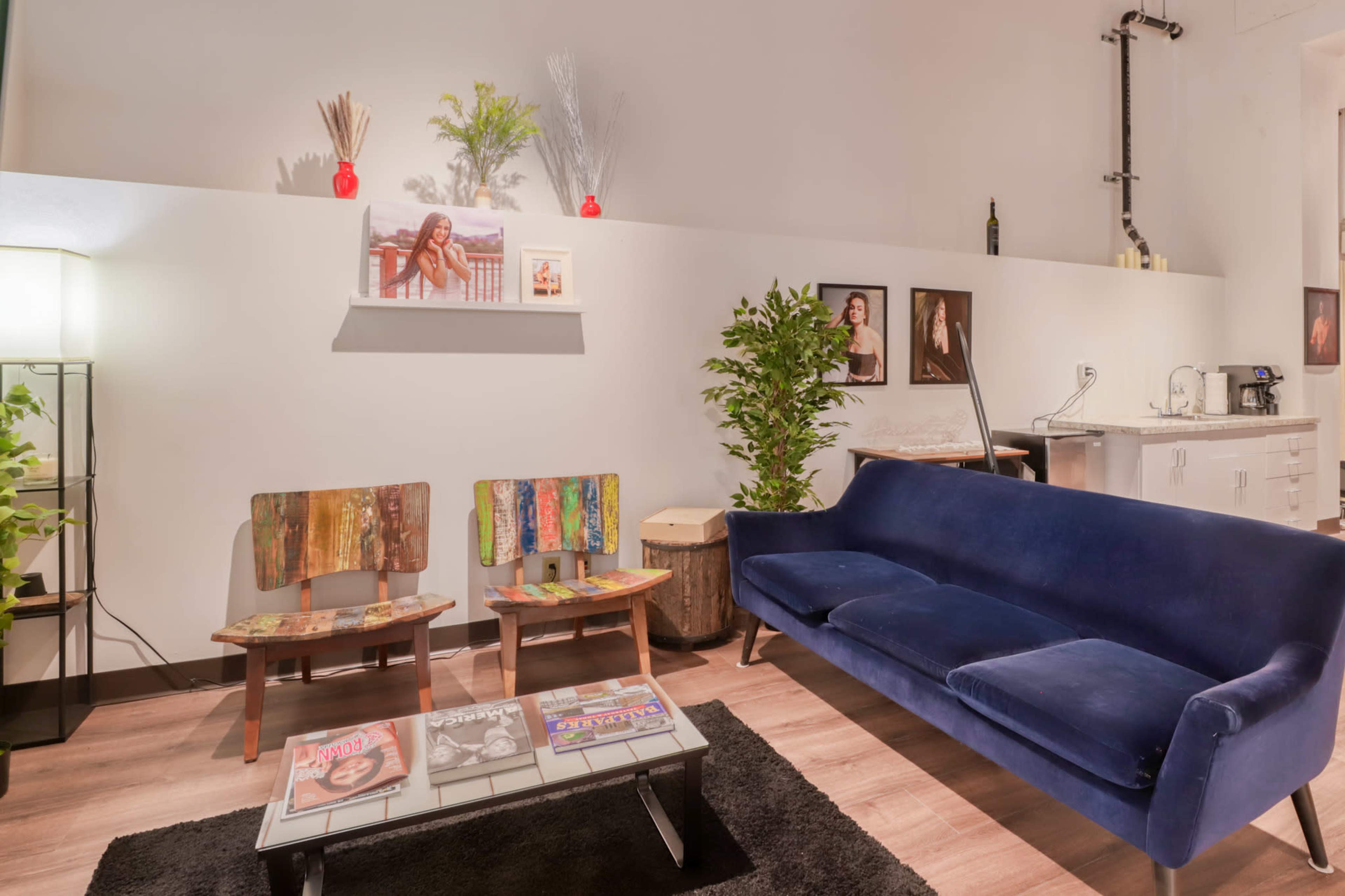 A modern living room features a blue sofa, two wooden chairs, a coffee table with magazines, and decorative plants against white walls adorned with framed photos.