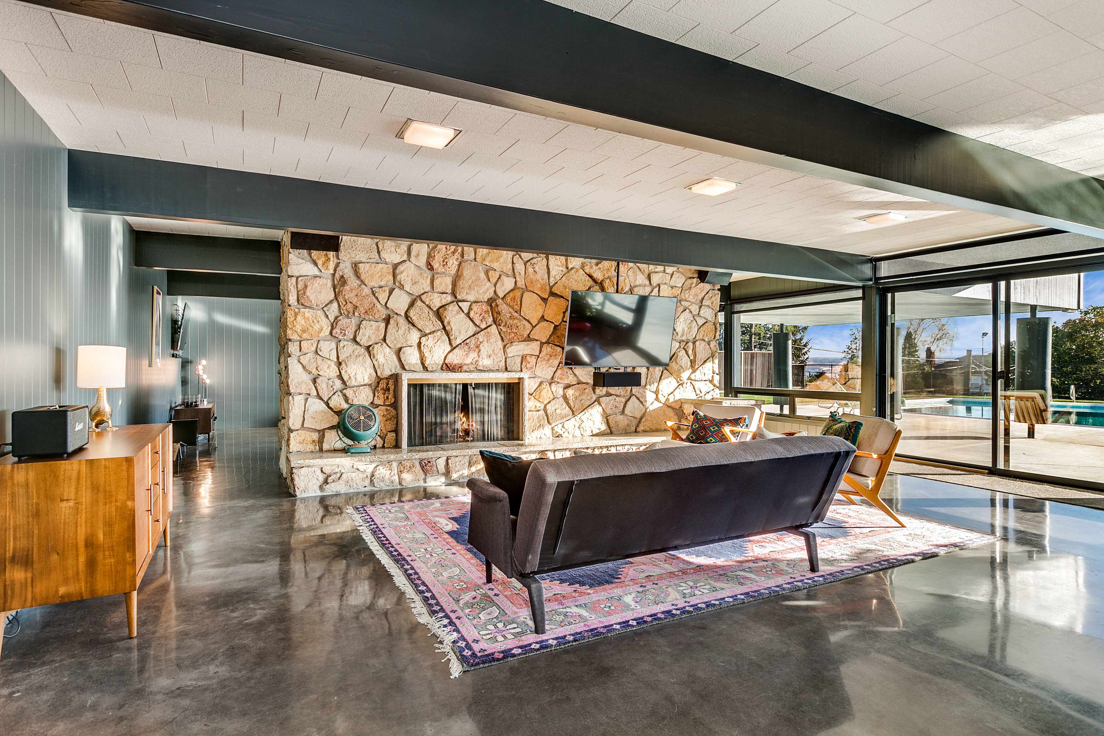 The living room features a stone fireplace, large glass doors, and modern furniture arranged on a colorful rug.