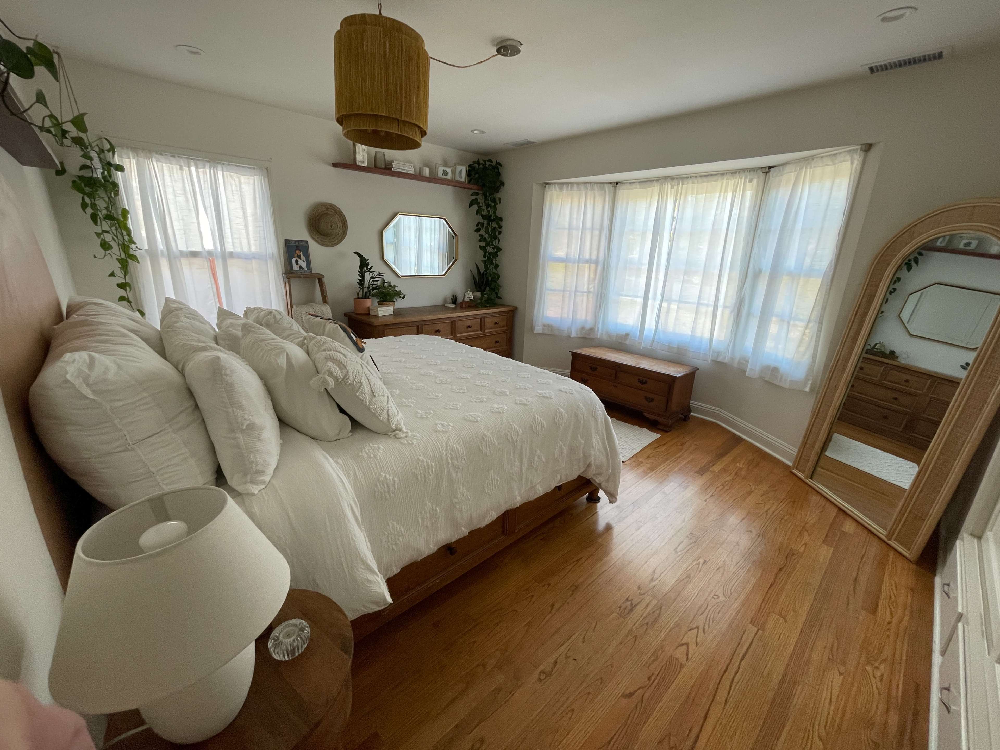 A well-furnished bedroom features a bed with white bedding, a wooden dresser, and large windows allowing natural light to fill the space.