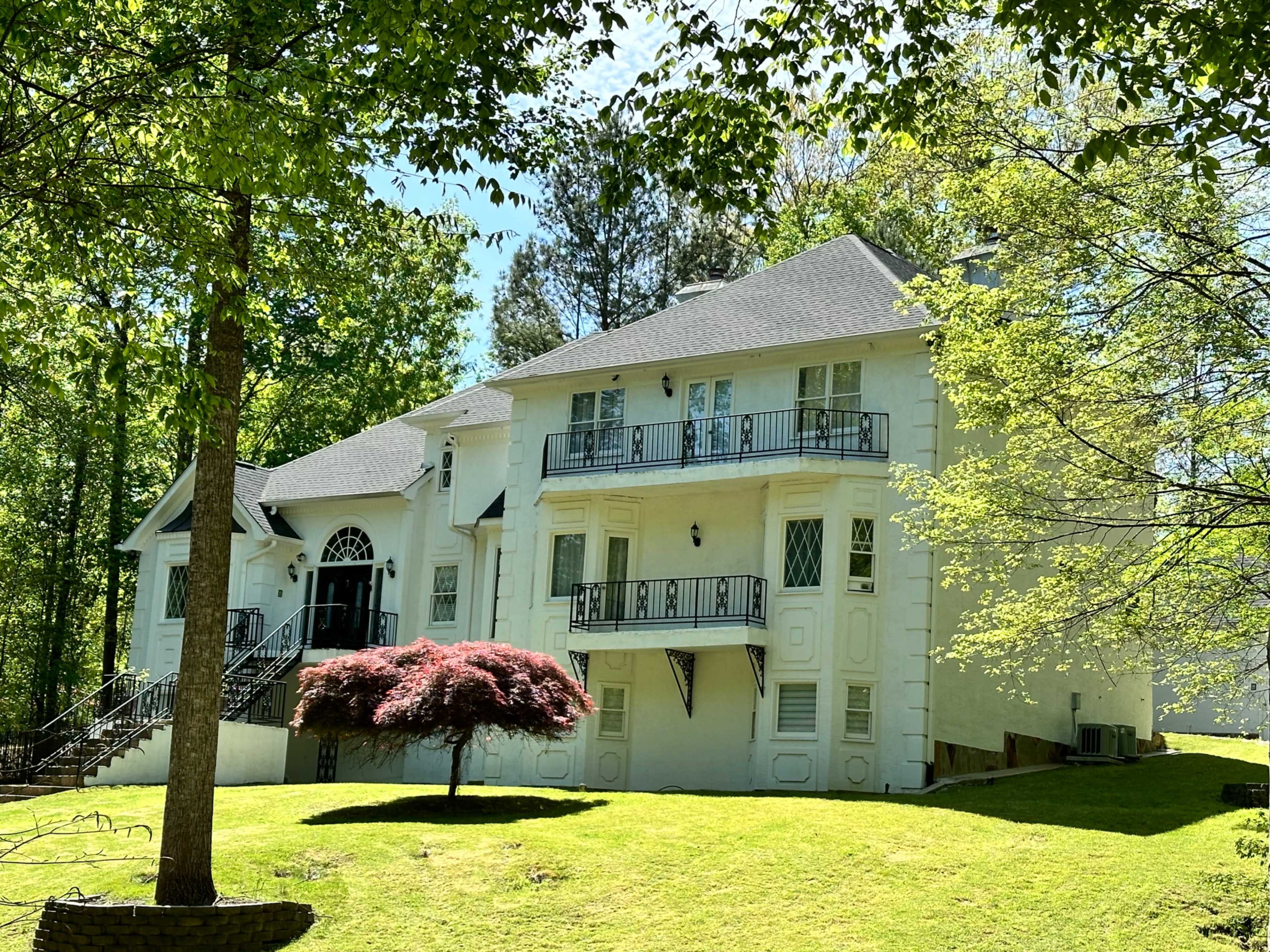 A two-story white house with a symmetrical facade and balconies is set against a backdrop of green trees and a manicured lawn.