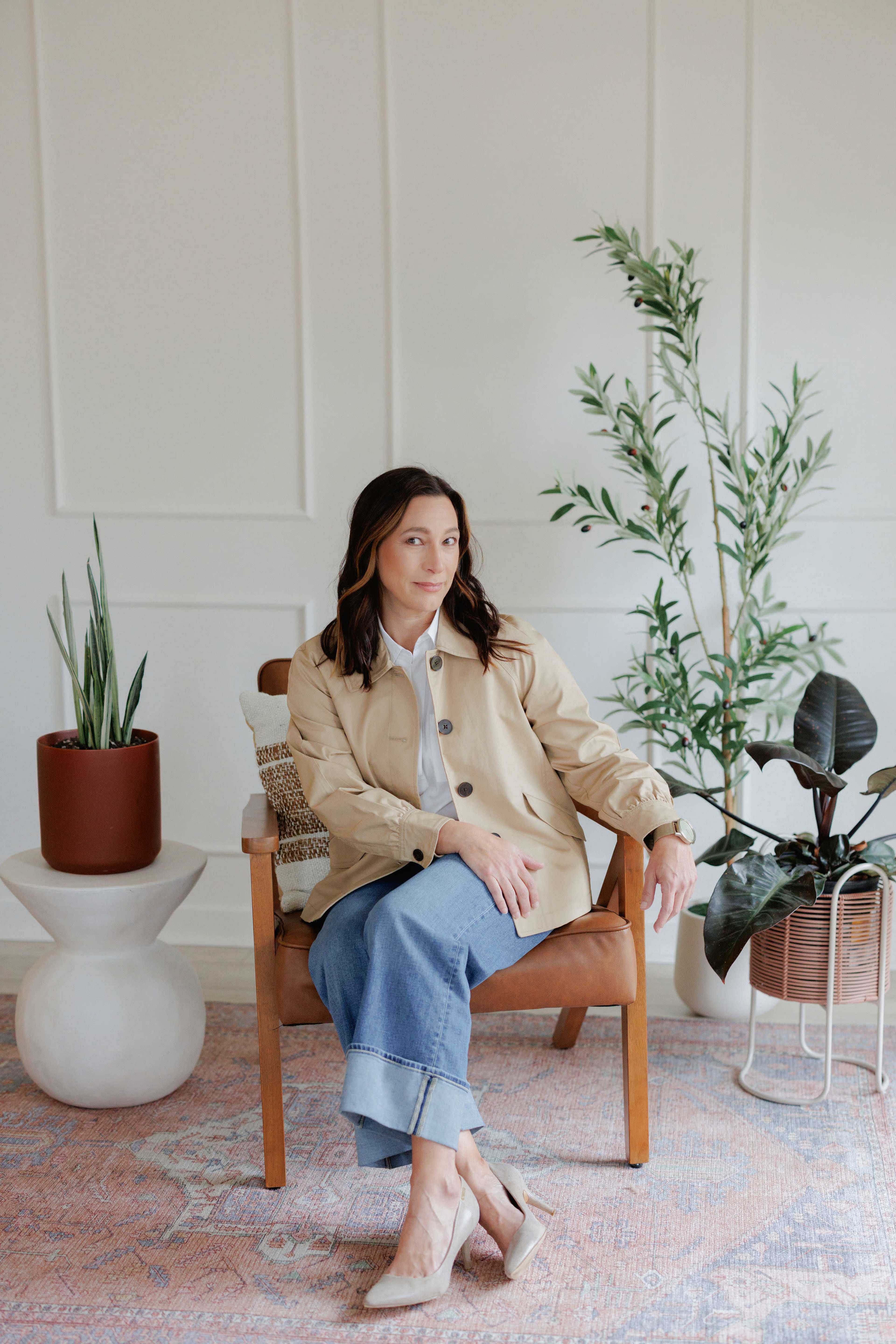 A woman in a beige jacket sits in a wooden chair in a room with indoor plants and a patterned rug.