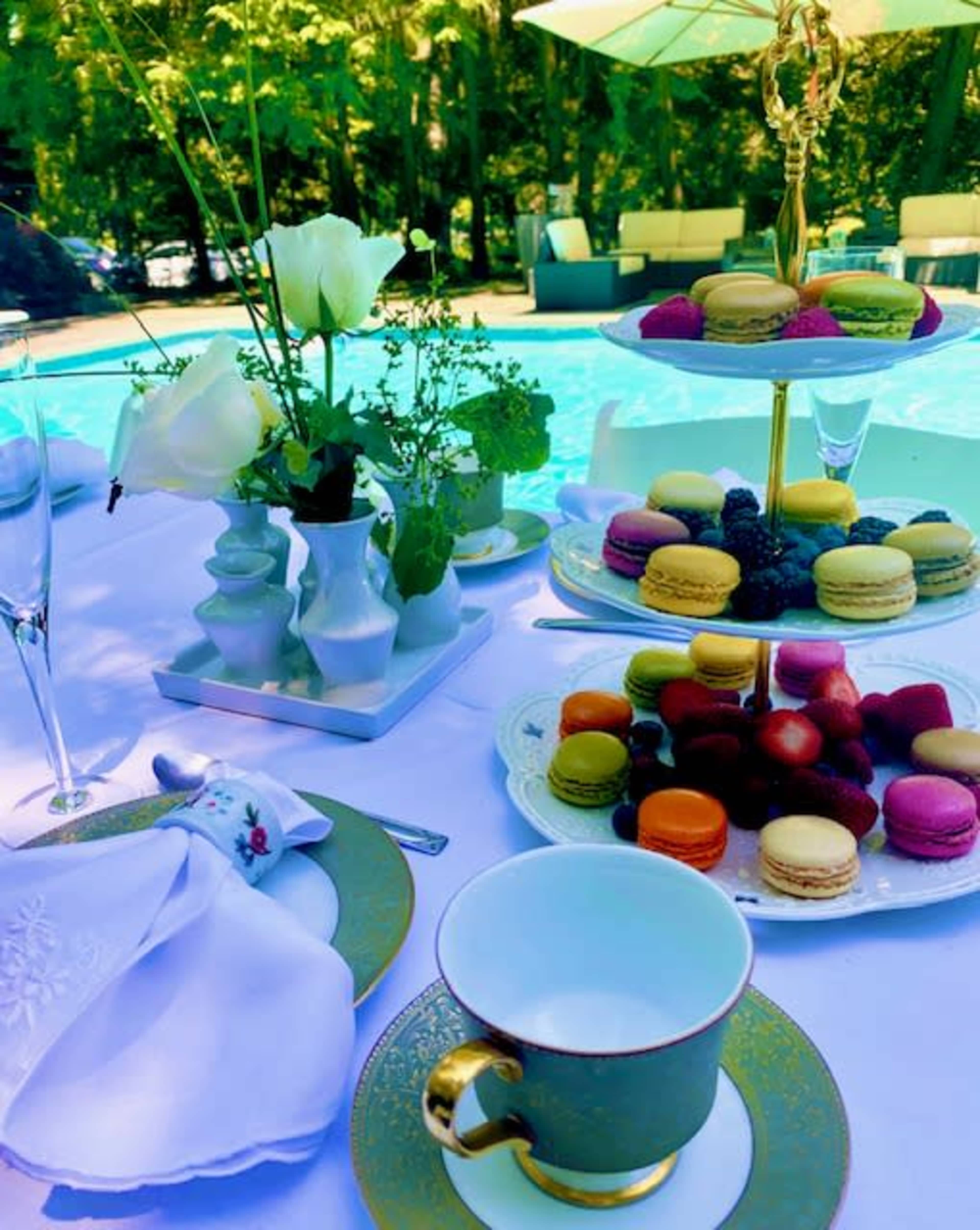 A table is set beside a pool, featuring an assortment of colorful macarons, fruit, and elegant tableware, along with a floral arrangement.