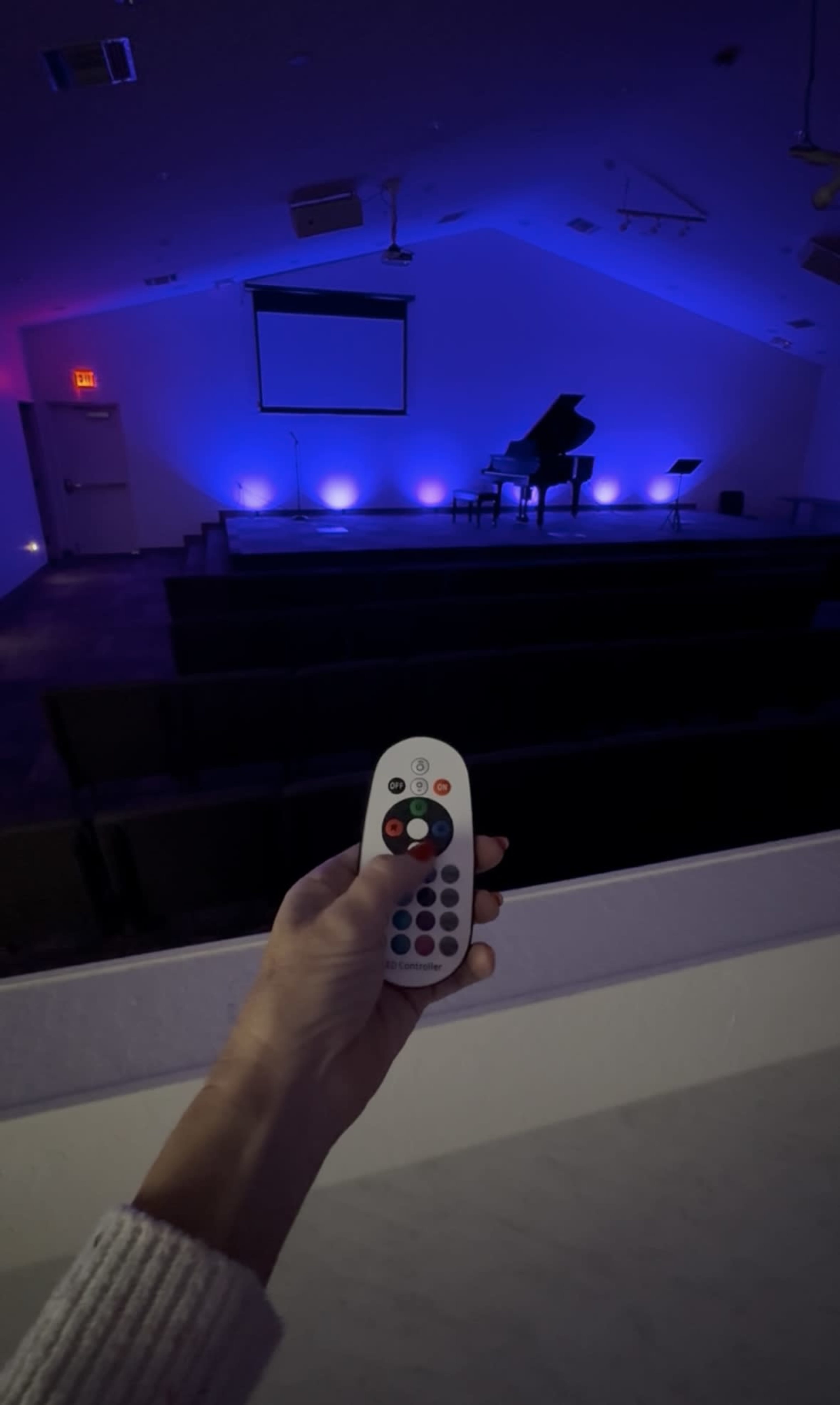 A person holds a remote control in front of a dimly lit stage featuring a grand piano and colorful lights.