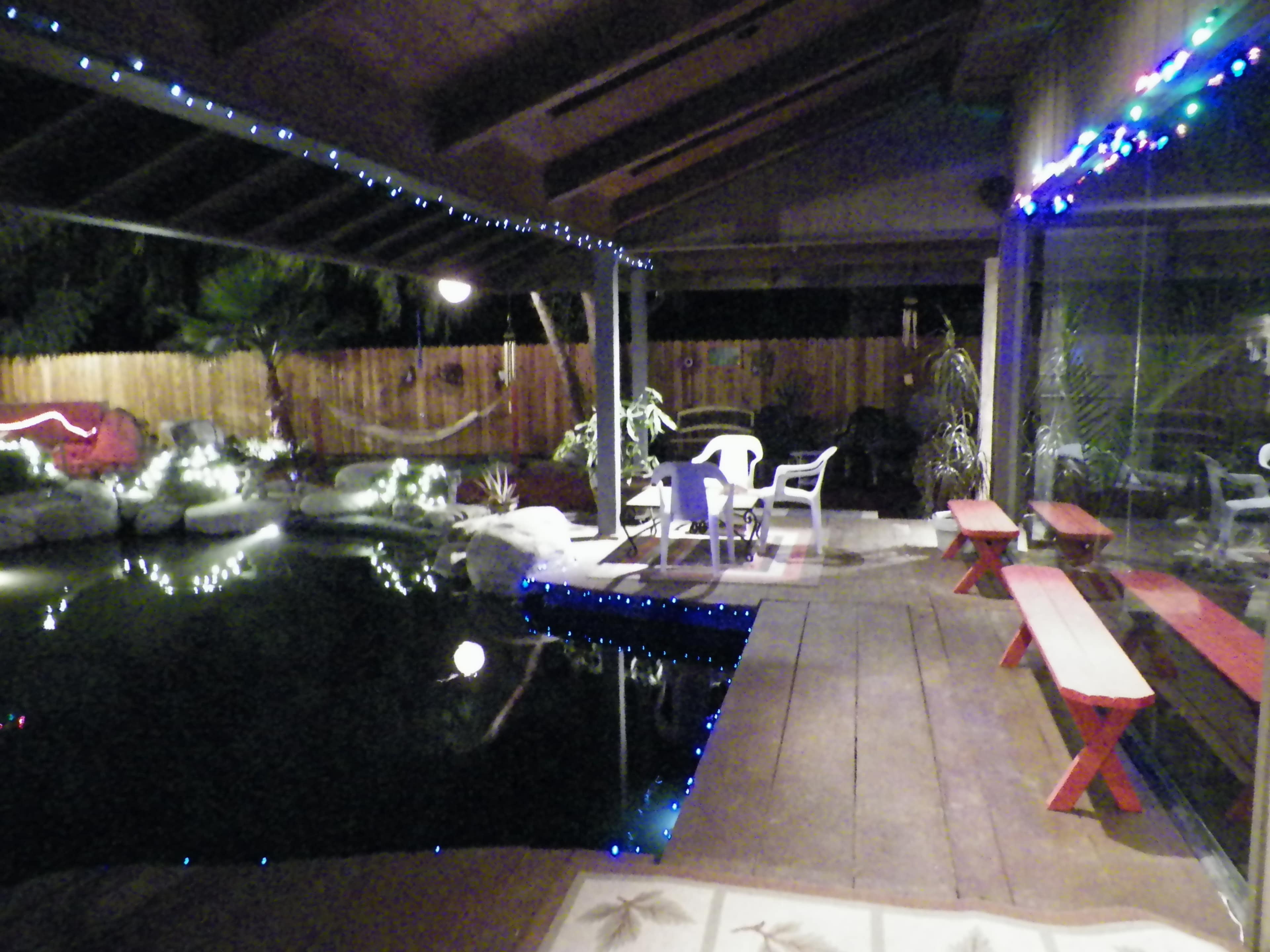 A backyard patio with a pool, featuring wooden benches, white chairs, and decorative lights along the structure.