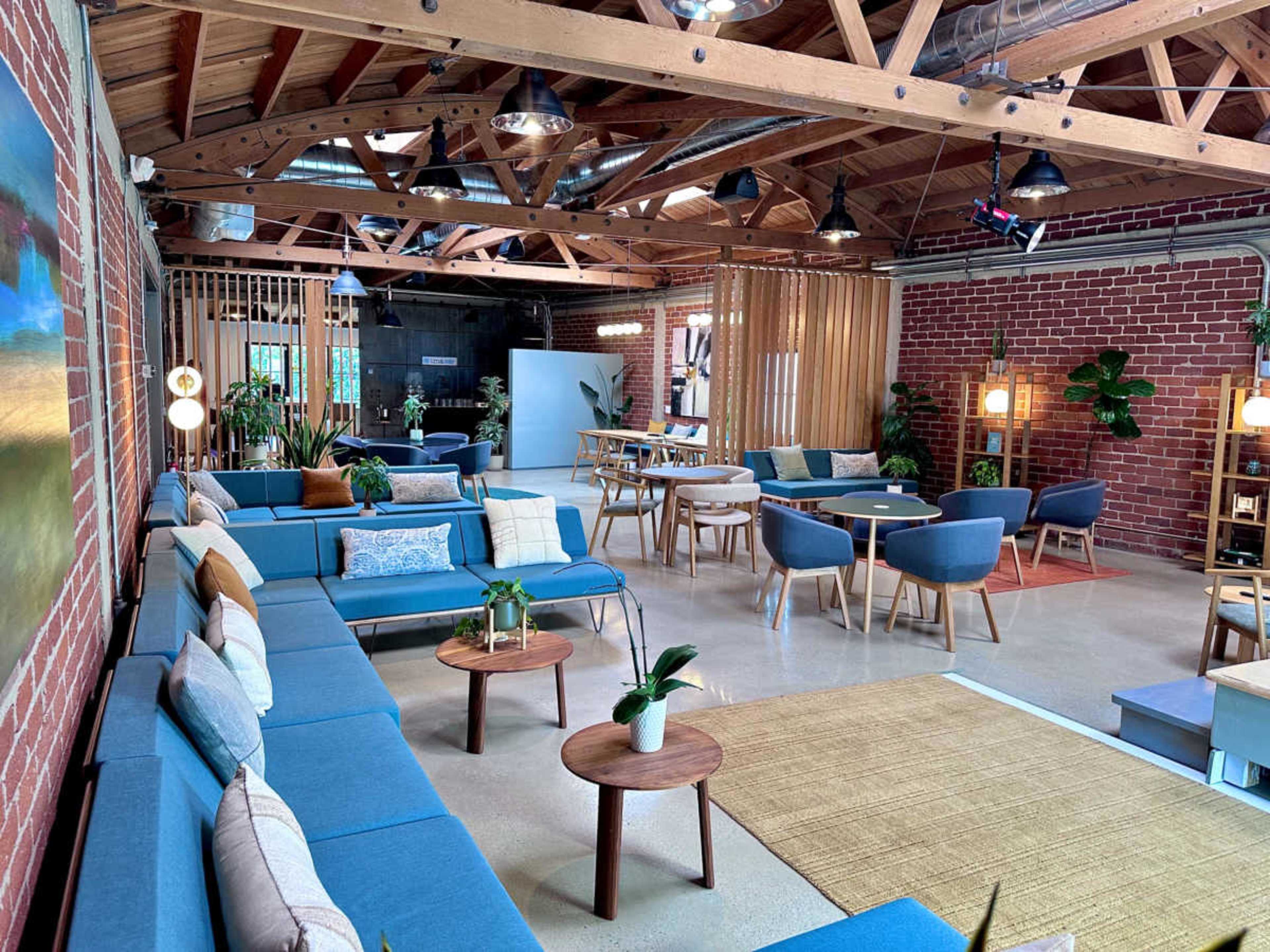 The image depicts a modern, open-plan lounge area with blue sofas, wooden tables, and plants, characterized by exposed brick walls and wooden beams.