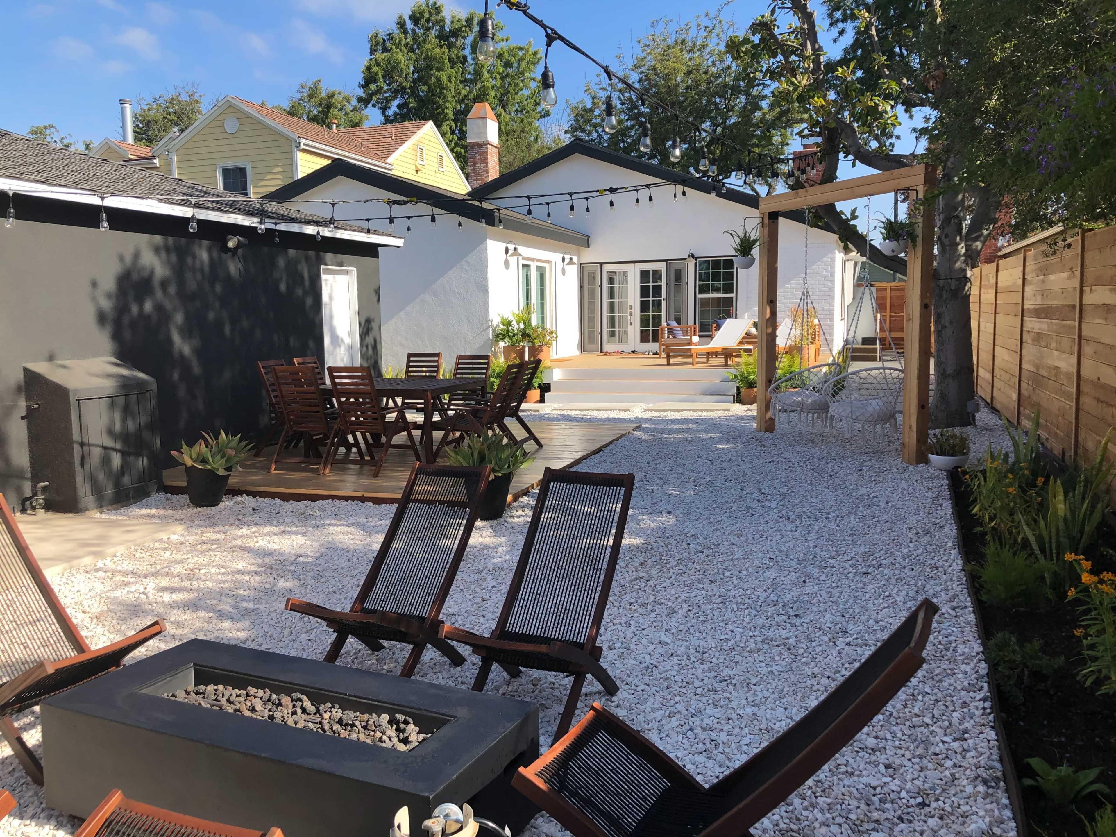 A backyard with a gravel surface, a seating area with wooden furniture, a fire pit, and a house with a patio in the background.