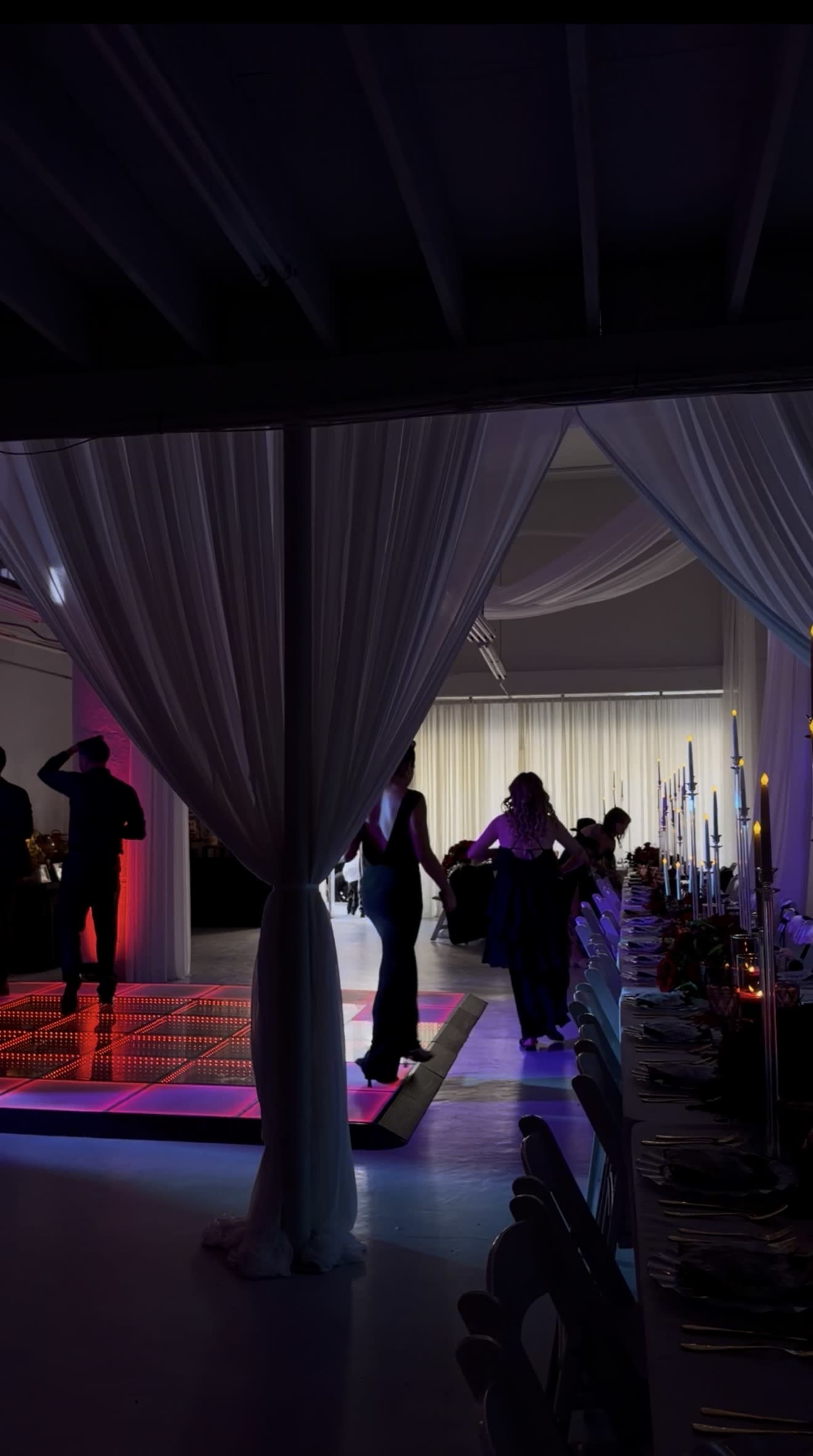 A brightly lit event space features a dance floor surrounded by elegantly set tables, with guests mingling in the background.