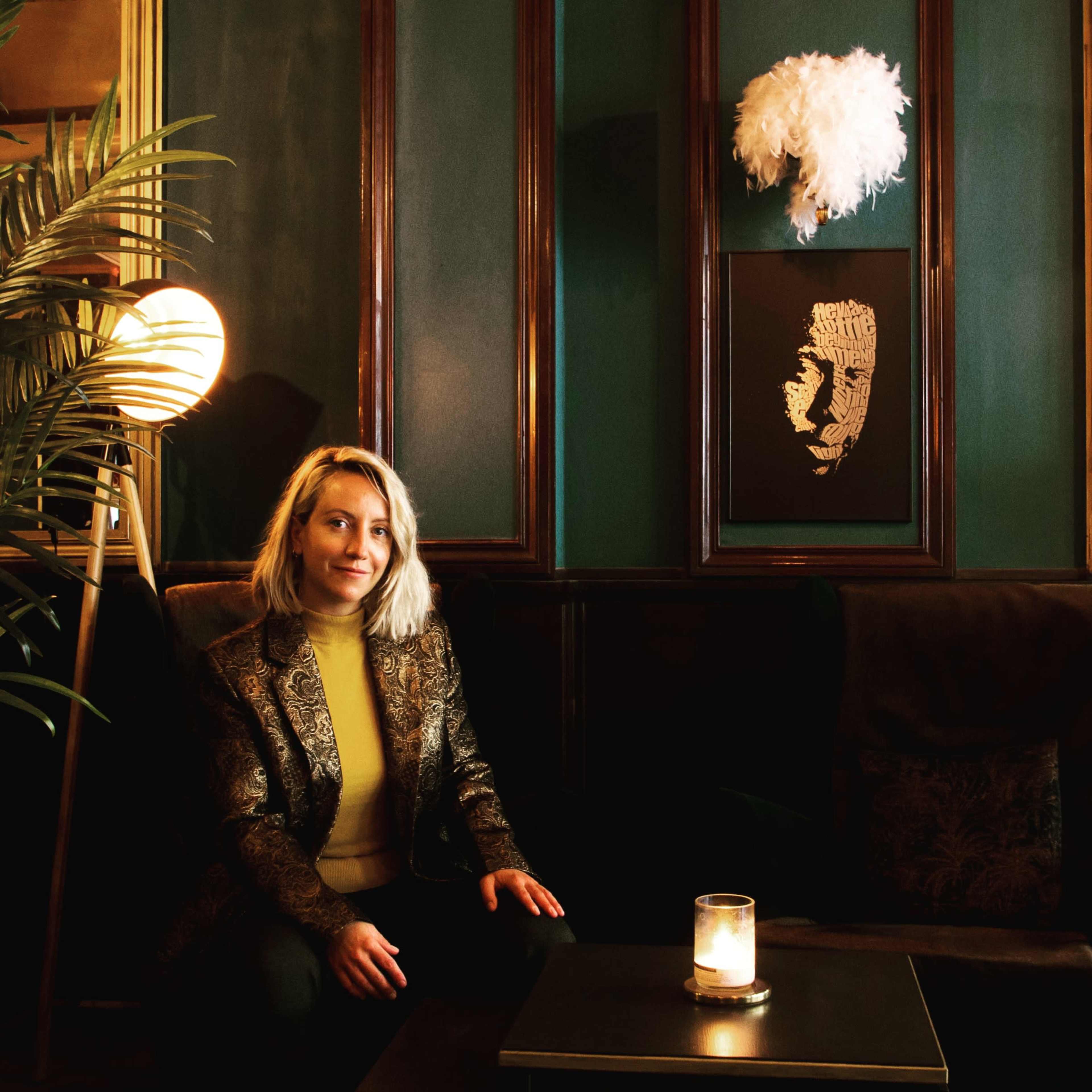 A woman with blonde hair sits at a table in a dimly lit, stylish interior featuring dark green walls and decorative artwork.