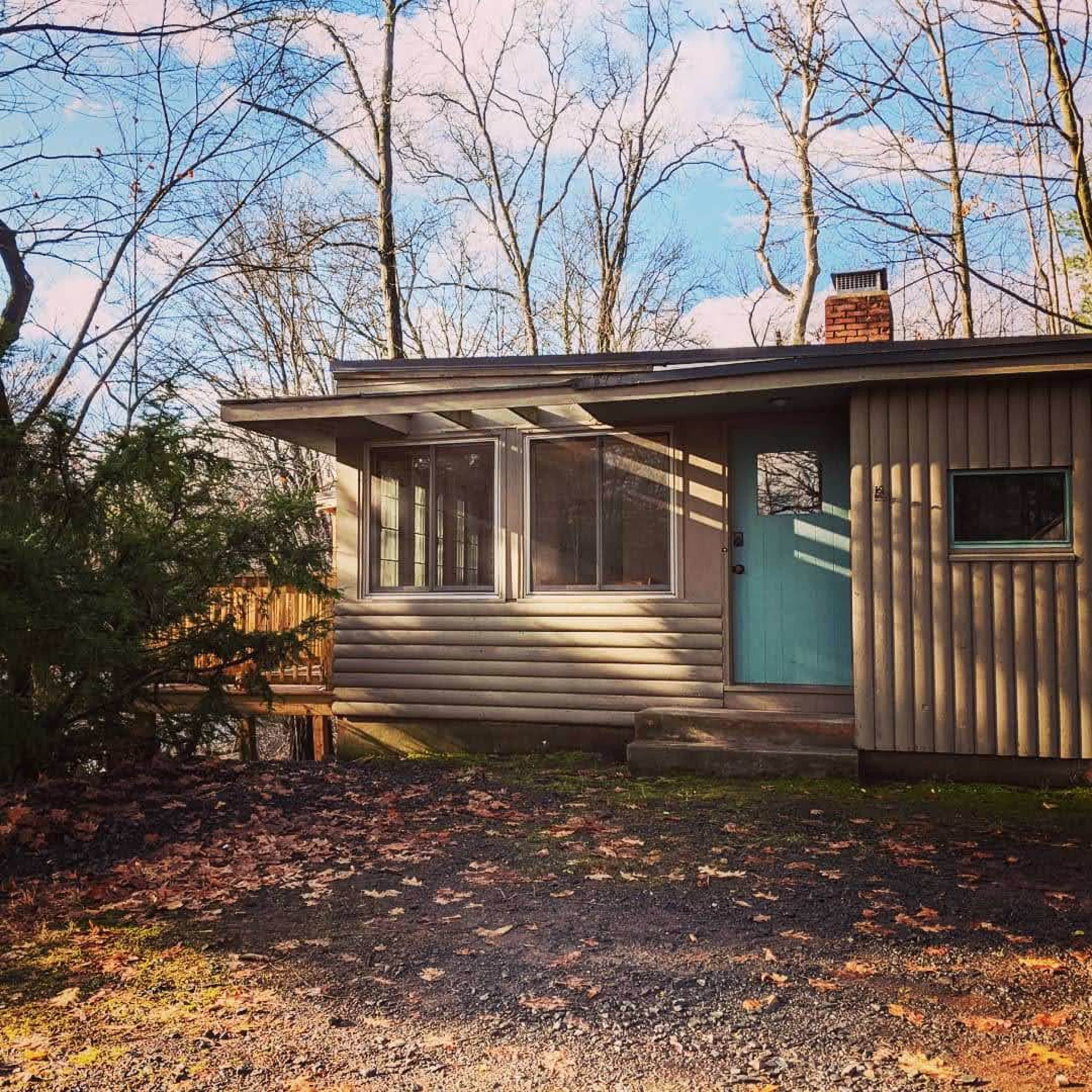 A small, modern house with a blue front door and large windows is surrounded by trees and bare branches.