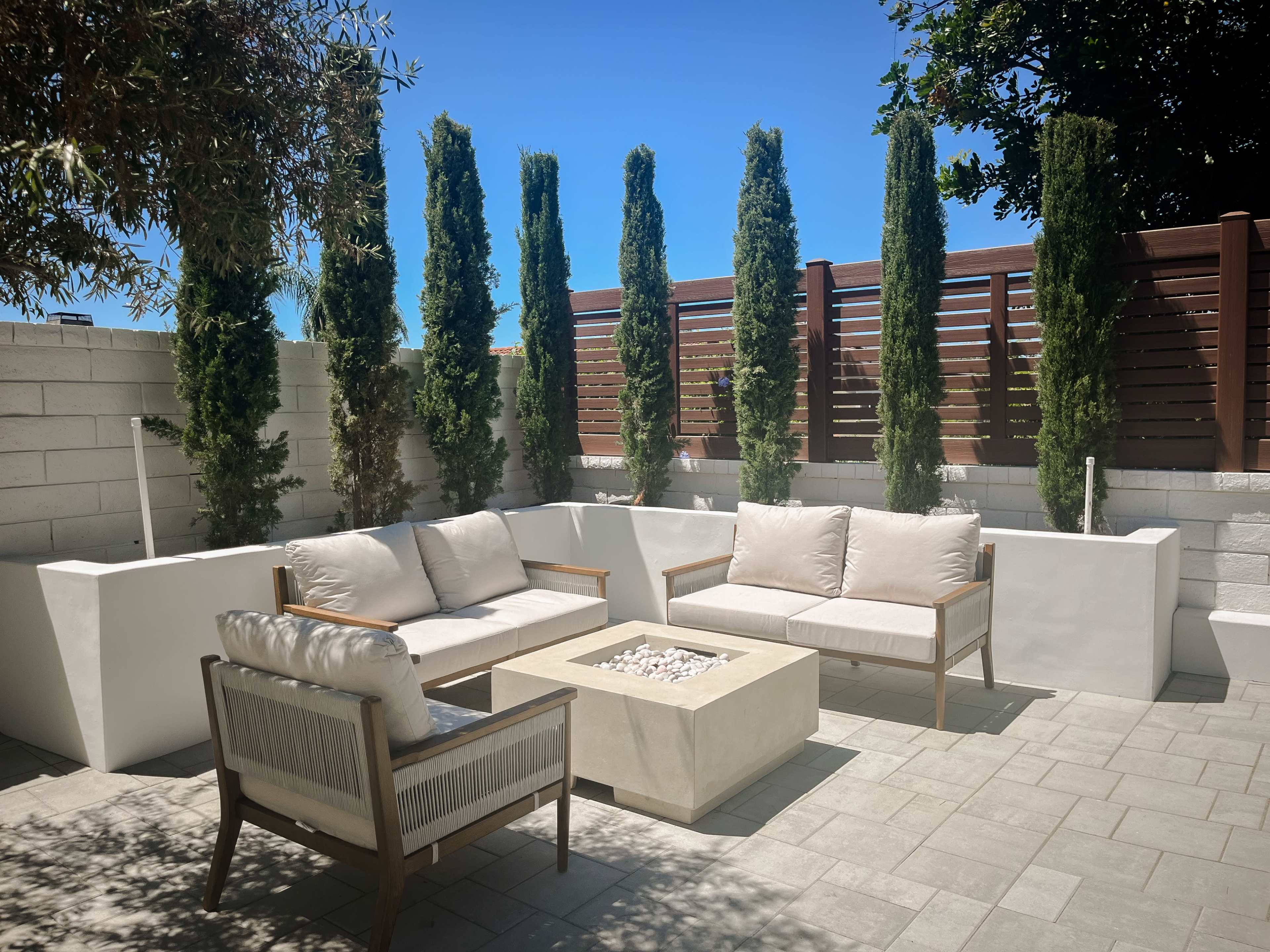 A modern outdoor seating area features light-colored furniture arranged around a square fire pit, surrounded by tall green trees and a privacy fence.