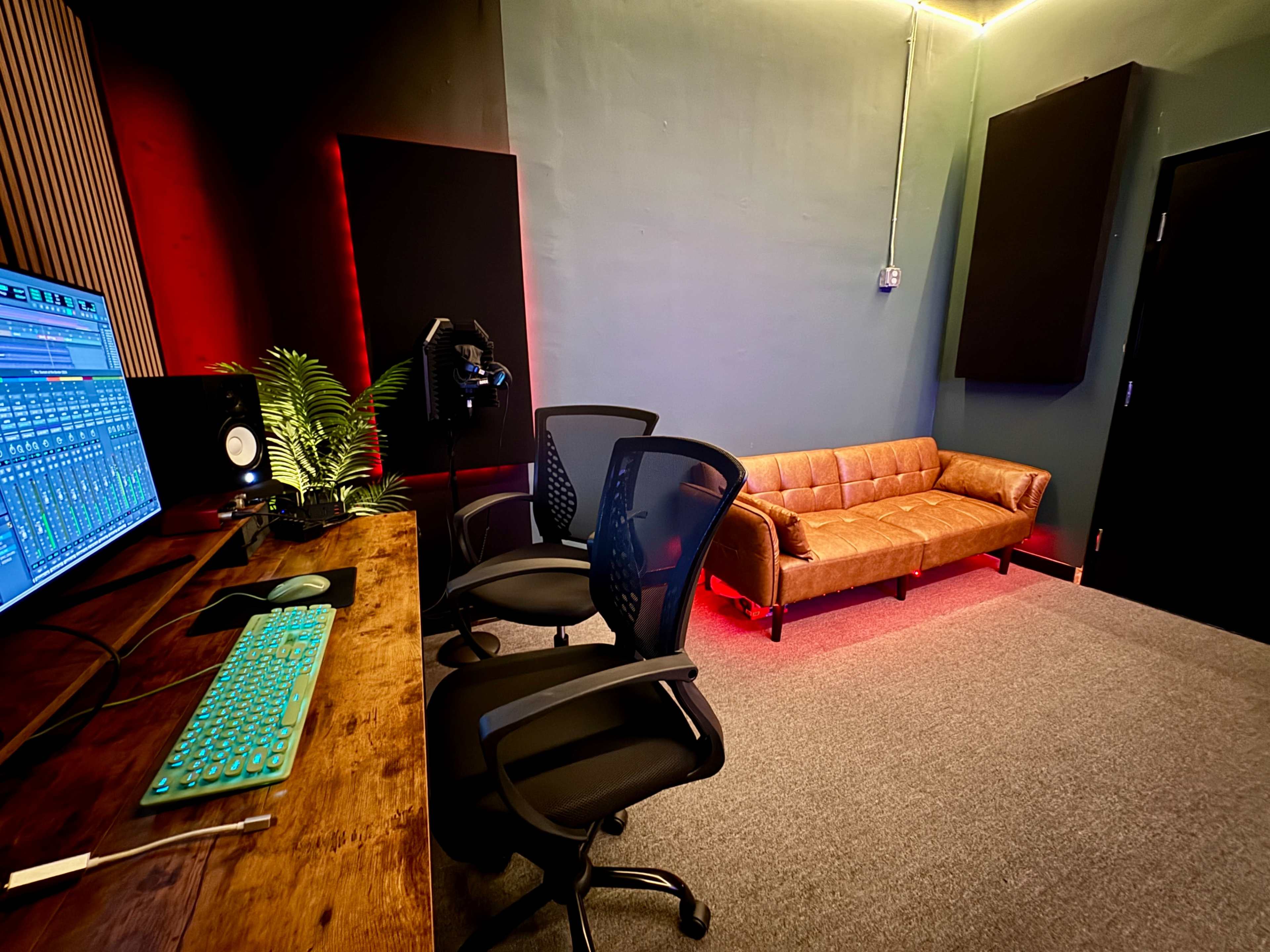 The image shows a modern audio recording studio with a wooden desk, two ergonomic chairs, a couch, and soundproof panels on the walls.
