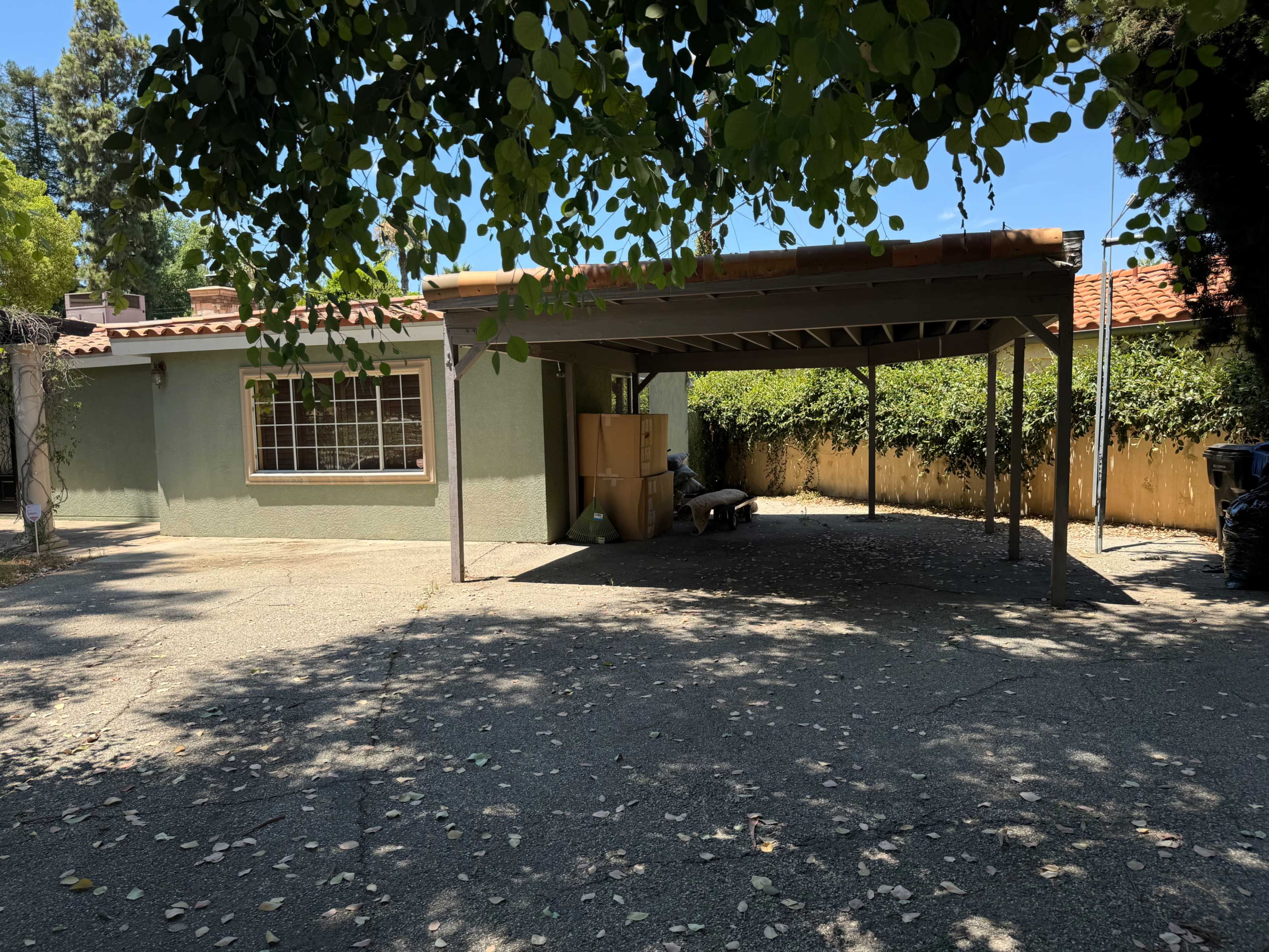 A carport shelters a portion of a house, surrounded by trees and a fenced yard.