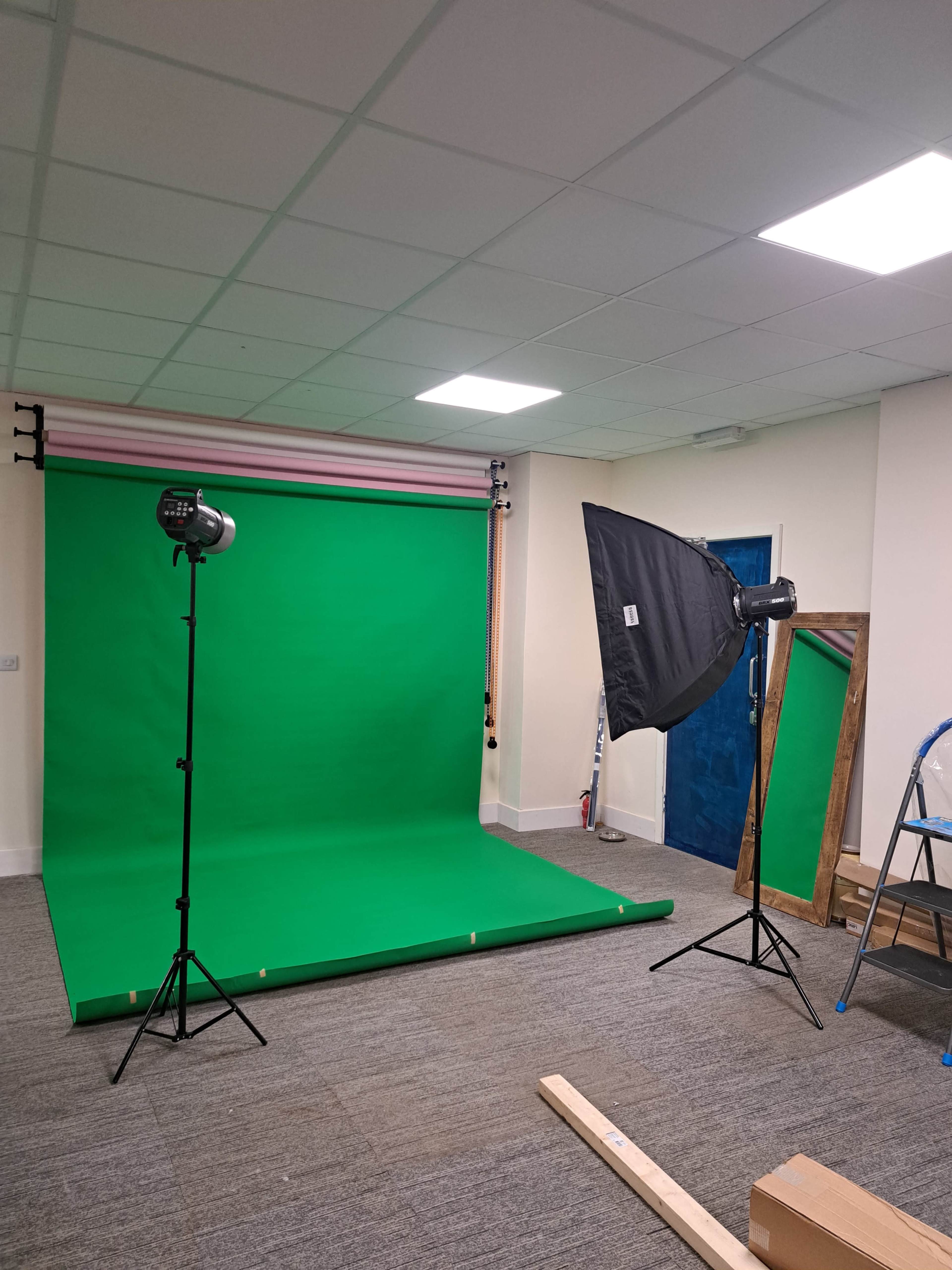 A photography studio setup with a green backdrop, two light stands, and a large mirror in a room with gray carpet.