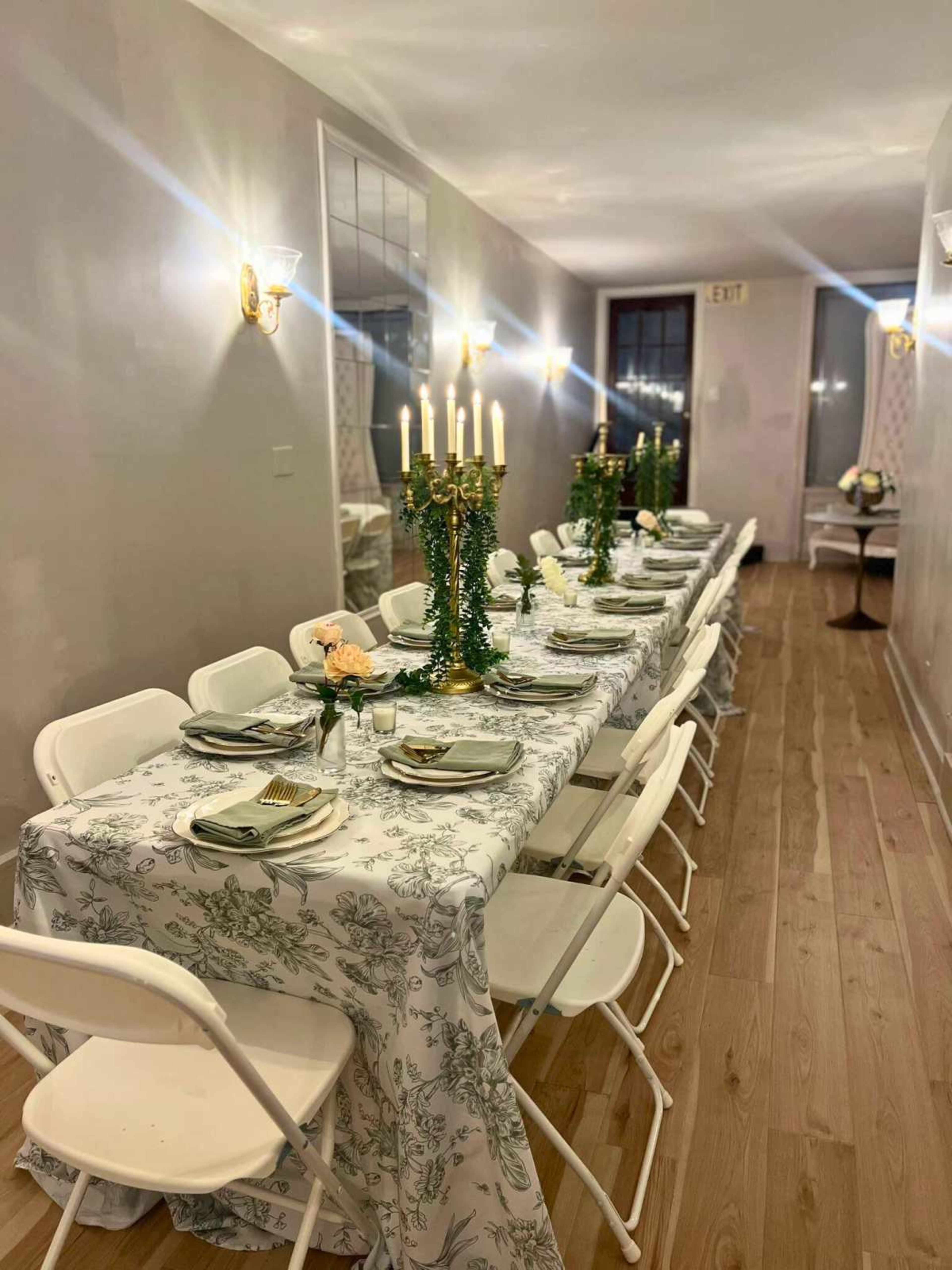 A long dining table is set with floral tablecloth, plates, and candelabras in a well-lit hallway.