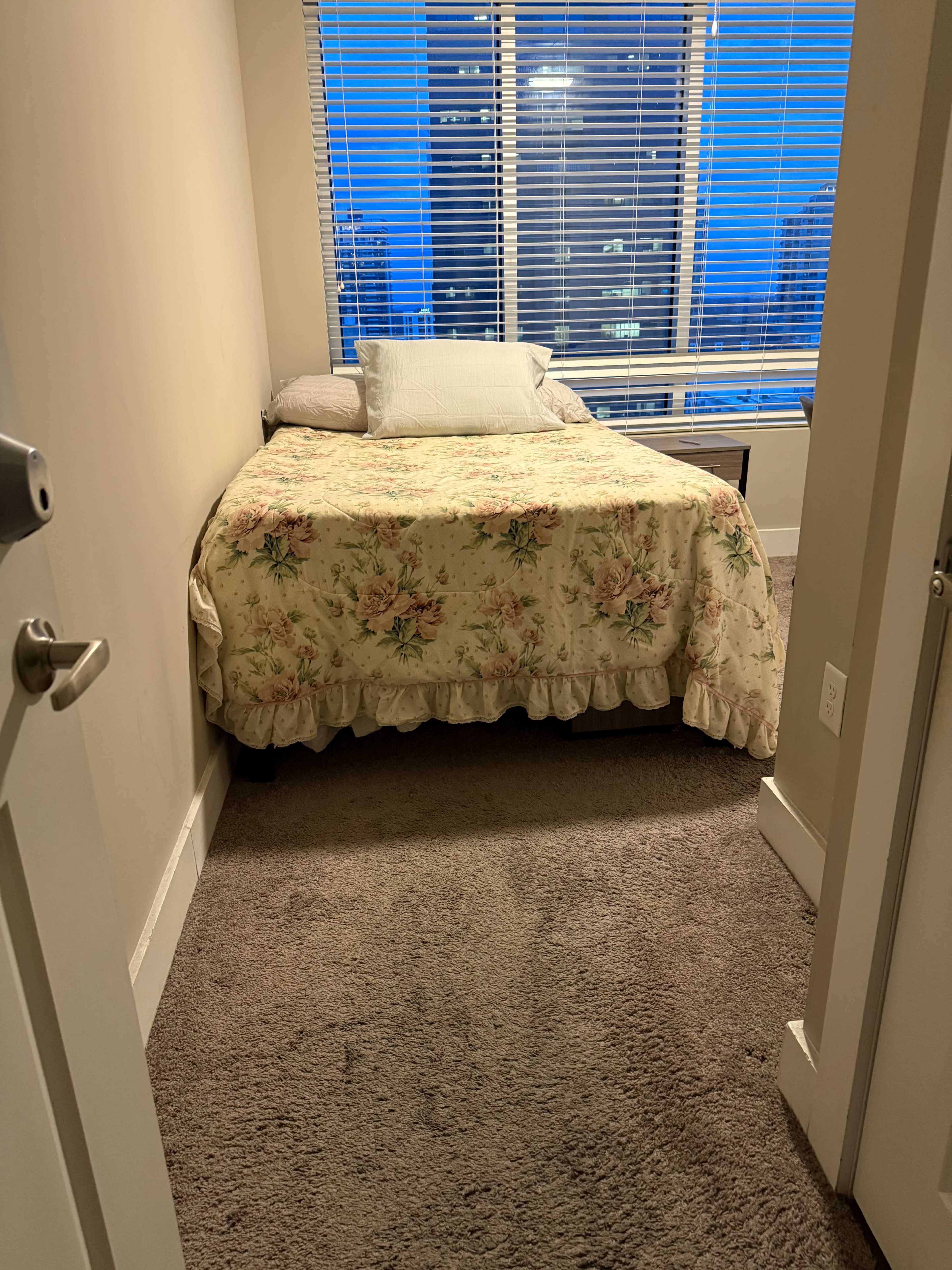 The image shows a small bedroom with a floral-patterned bedspread, a pillow, and large windows with blinds letting in evening light.