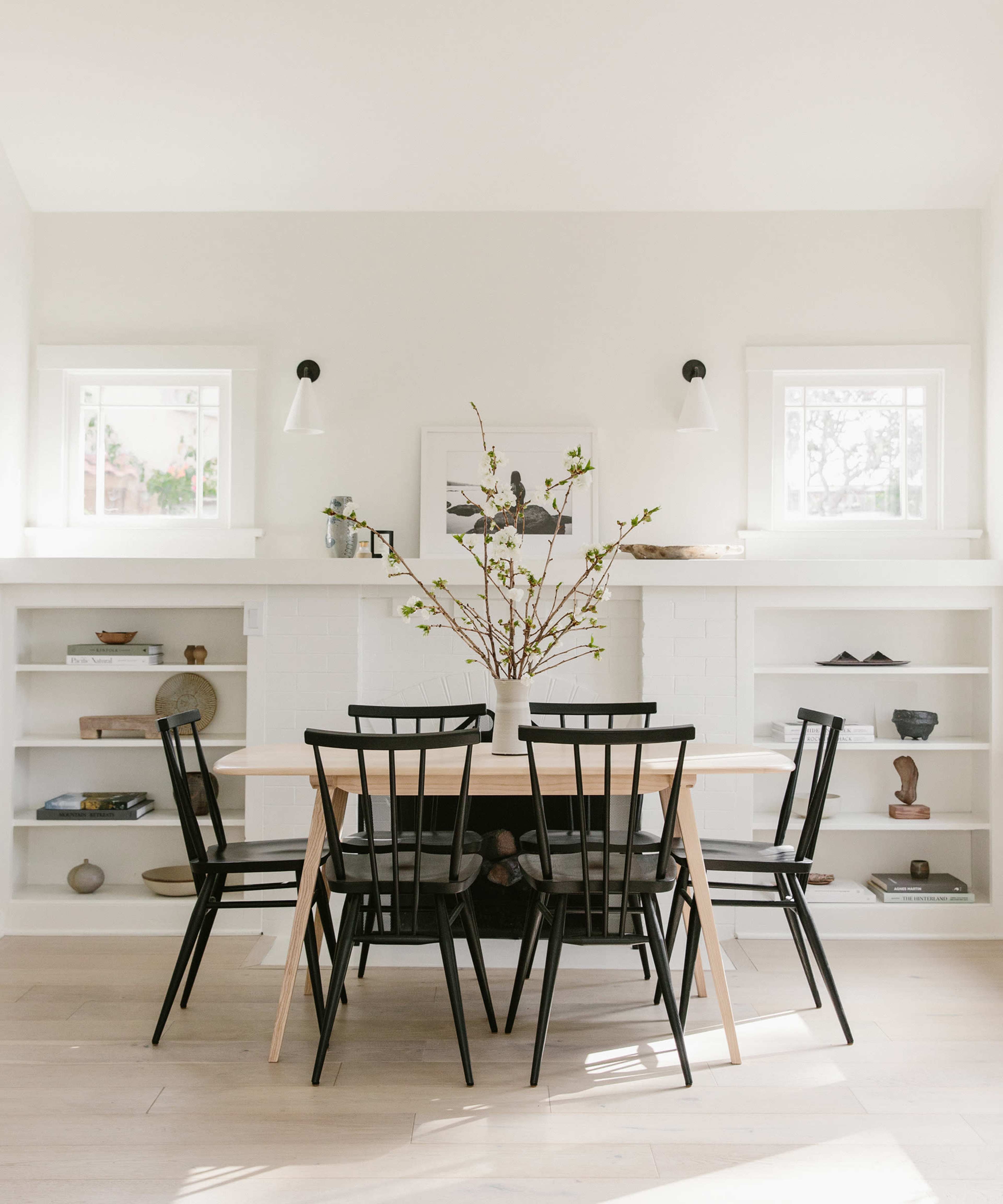 A simple dining area features a wooden table surrounded by black chairs, with a branch centerpiece and shelves displaying various decorative objects.