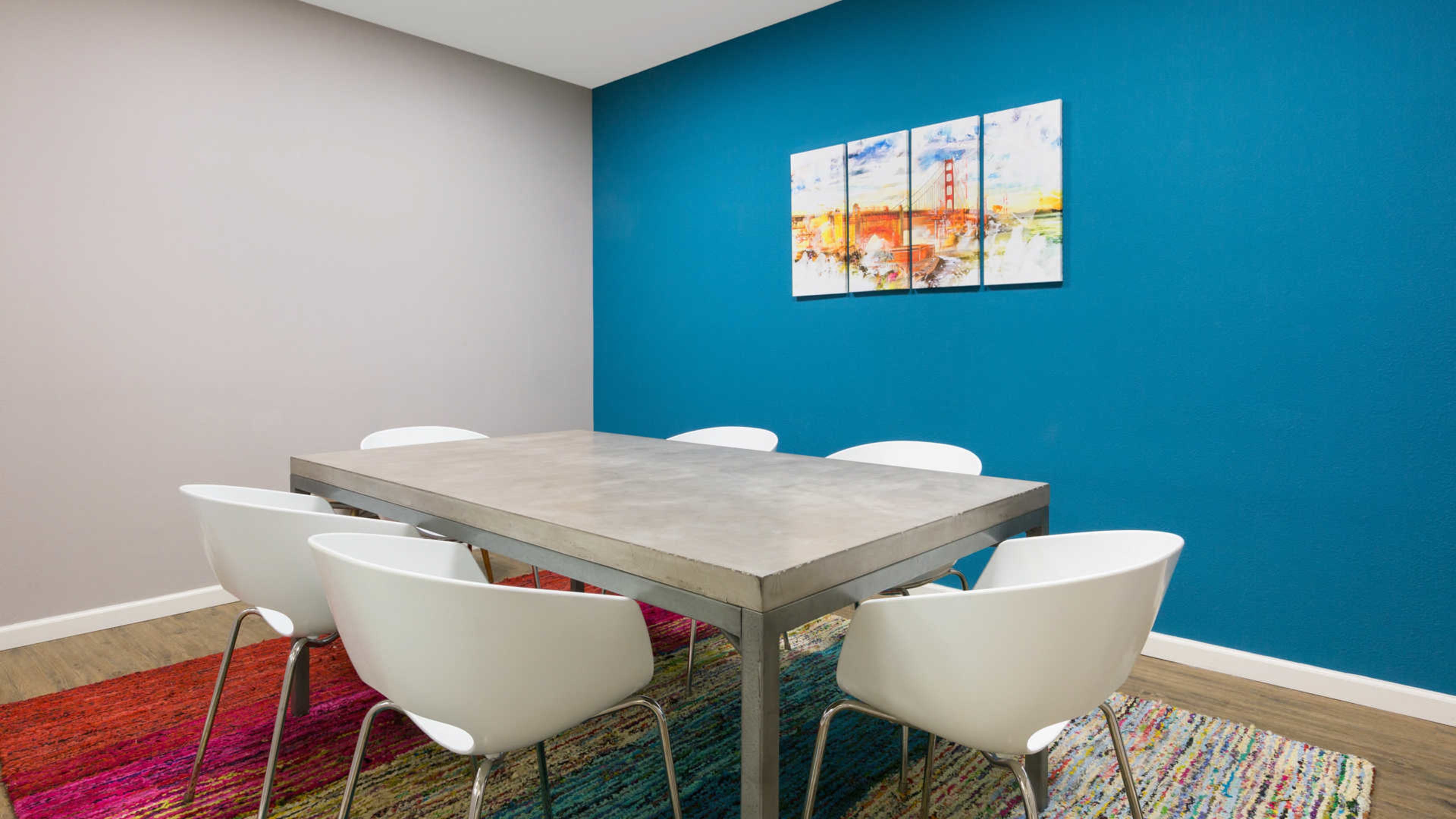 A modern dining area features a metallic table surrounded by six white chairs, with a colorful rug and a landscape painting on the blue wall.