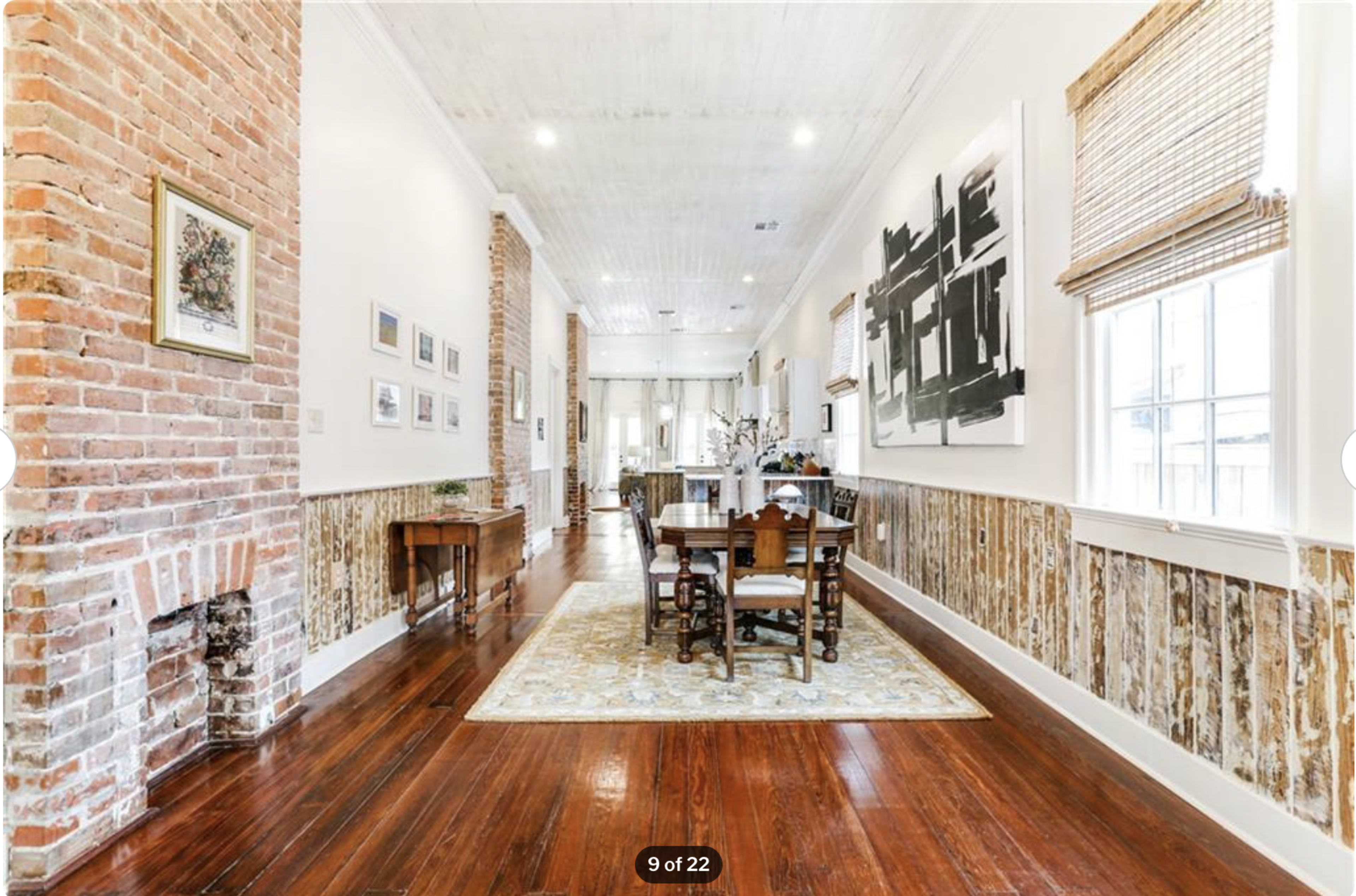 A long, narrow dining area with exposed brick walls, wooden flooring, and a table surrounded by chairs, leading toward a well-lit space with large windows.
