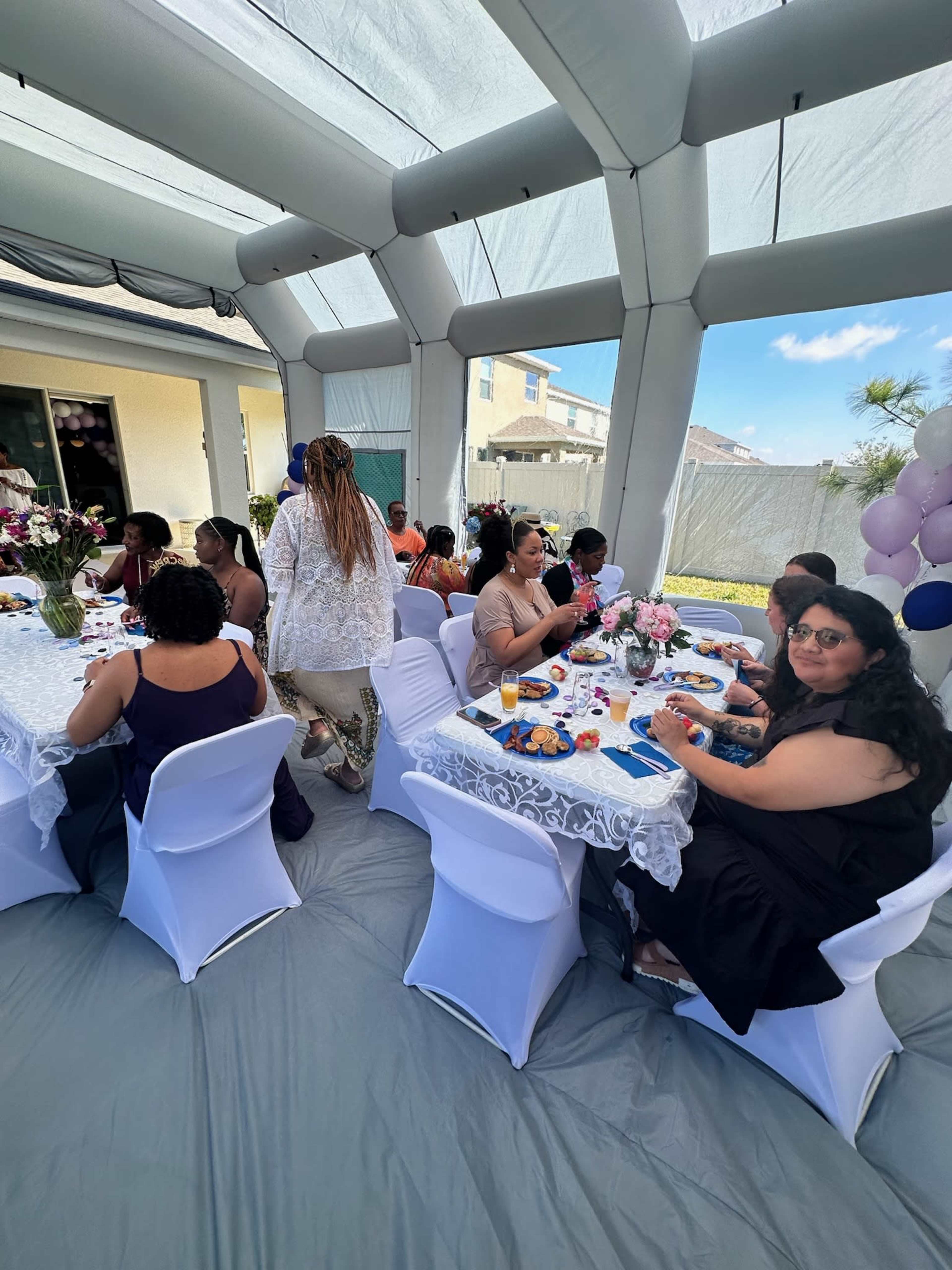 A group of people are seated at tables under a white tent, enjoying food and drinks during a gathering.