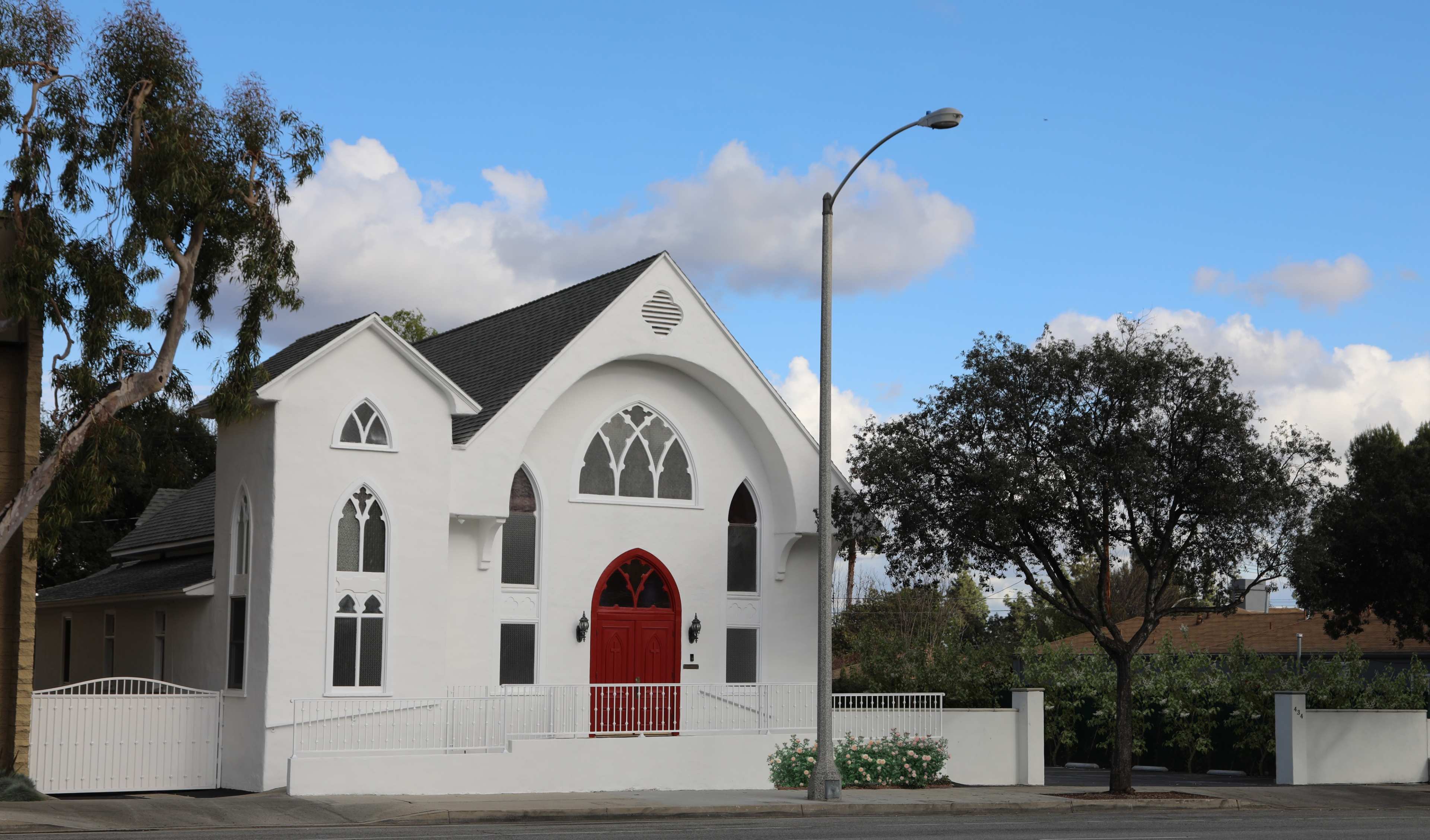 A white building with arched windows and a red door stands on a quiet street under a blue sky with scattered clouds.