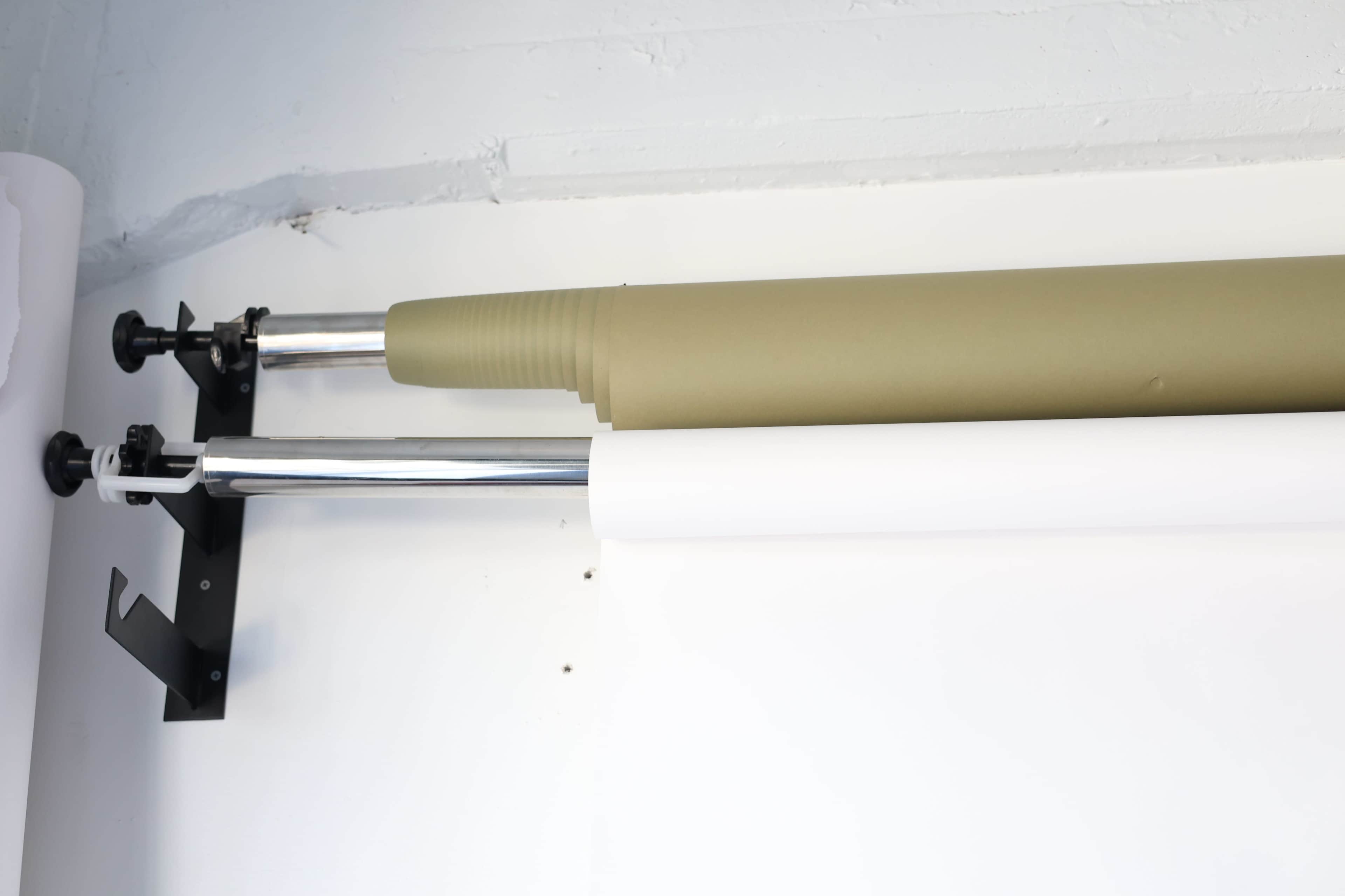 Two rolls of backdrop paper, one olive green and one white, are mounted on metal poles against a white wall.