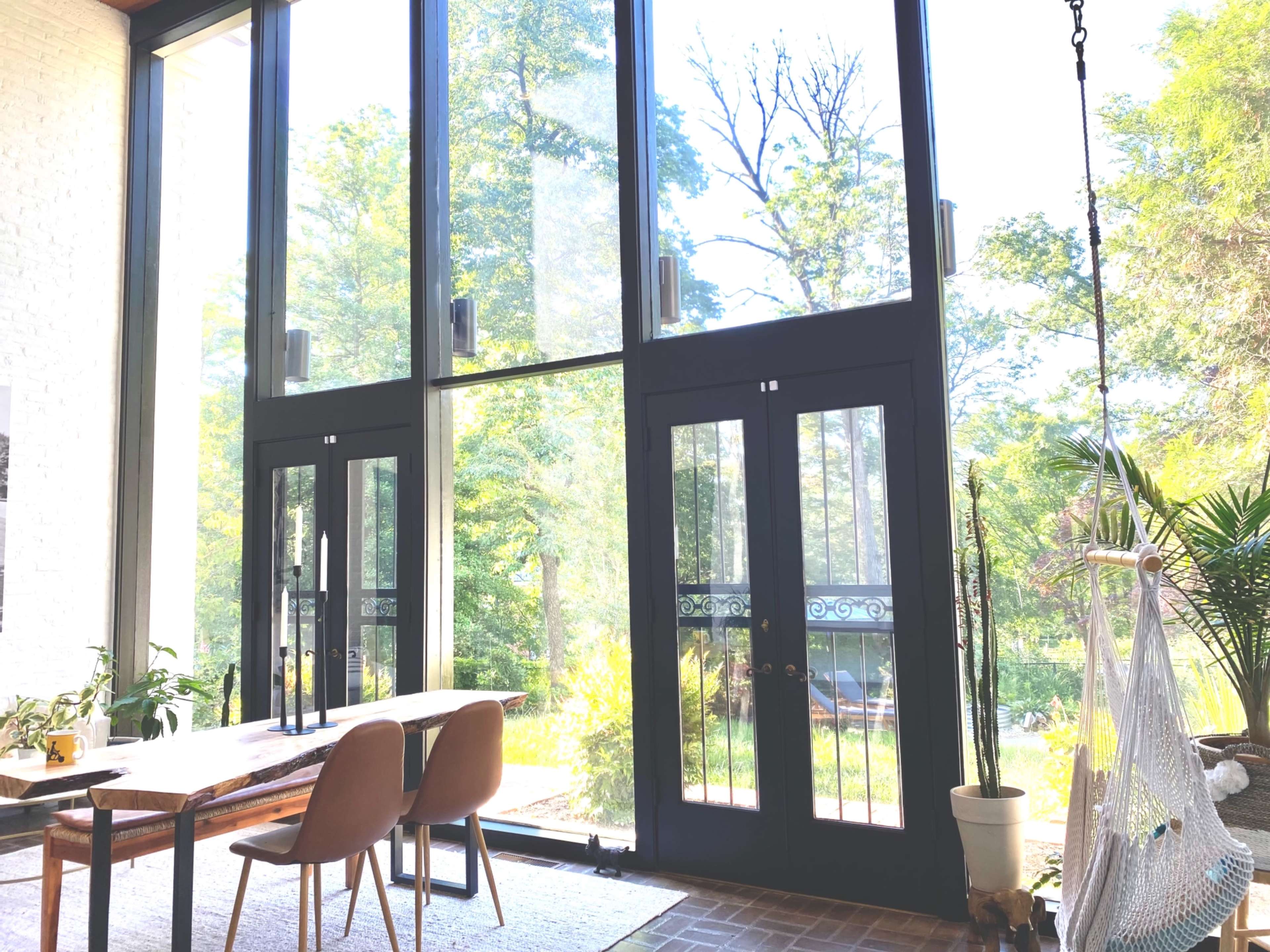 A sunlit room features large glass doors, a wooden table with two chairs, and a hammock in a corner, surrounded by greenery.