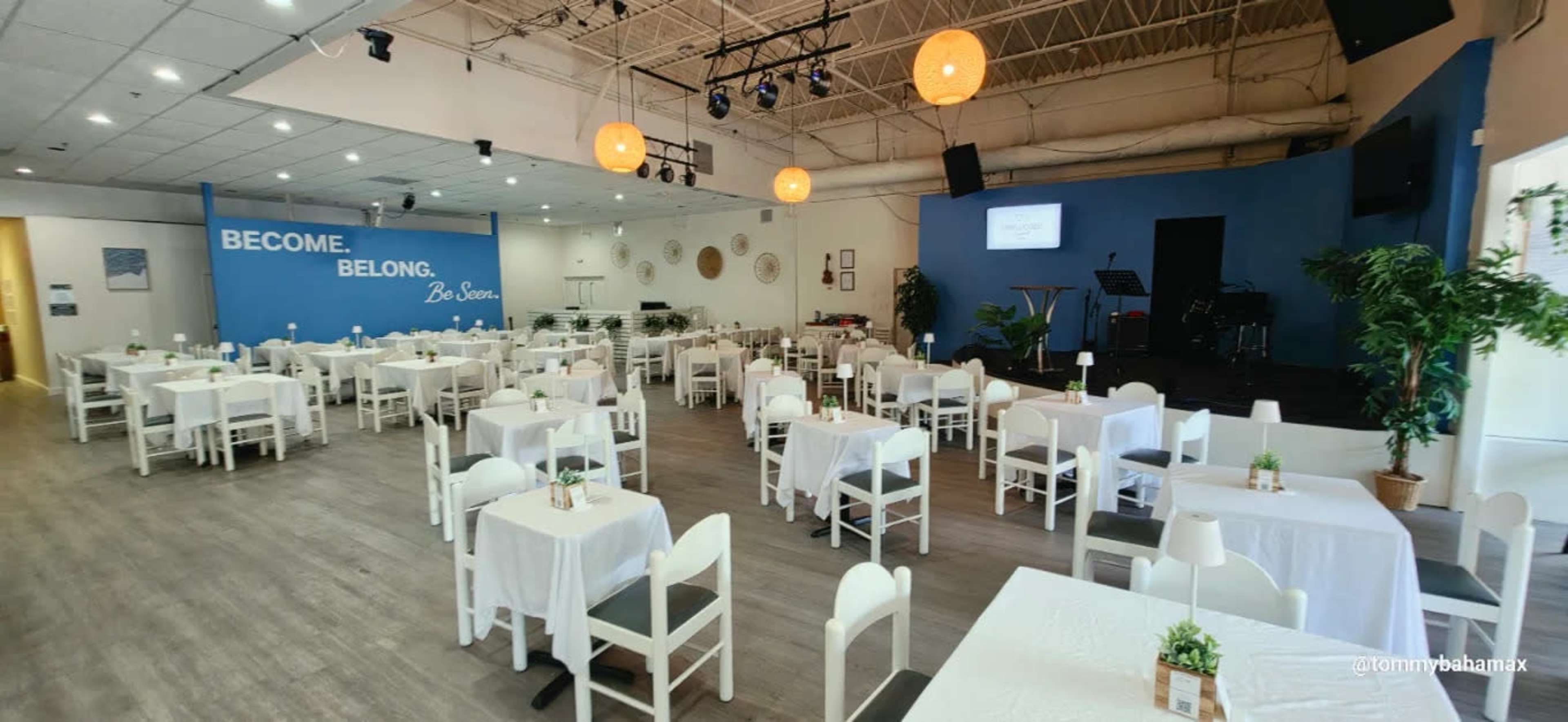 The image shows a spacious event venue set up with white tables and chairs, decorated with small centerpieces, and features a stage area with musical instruments against a blue wall.