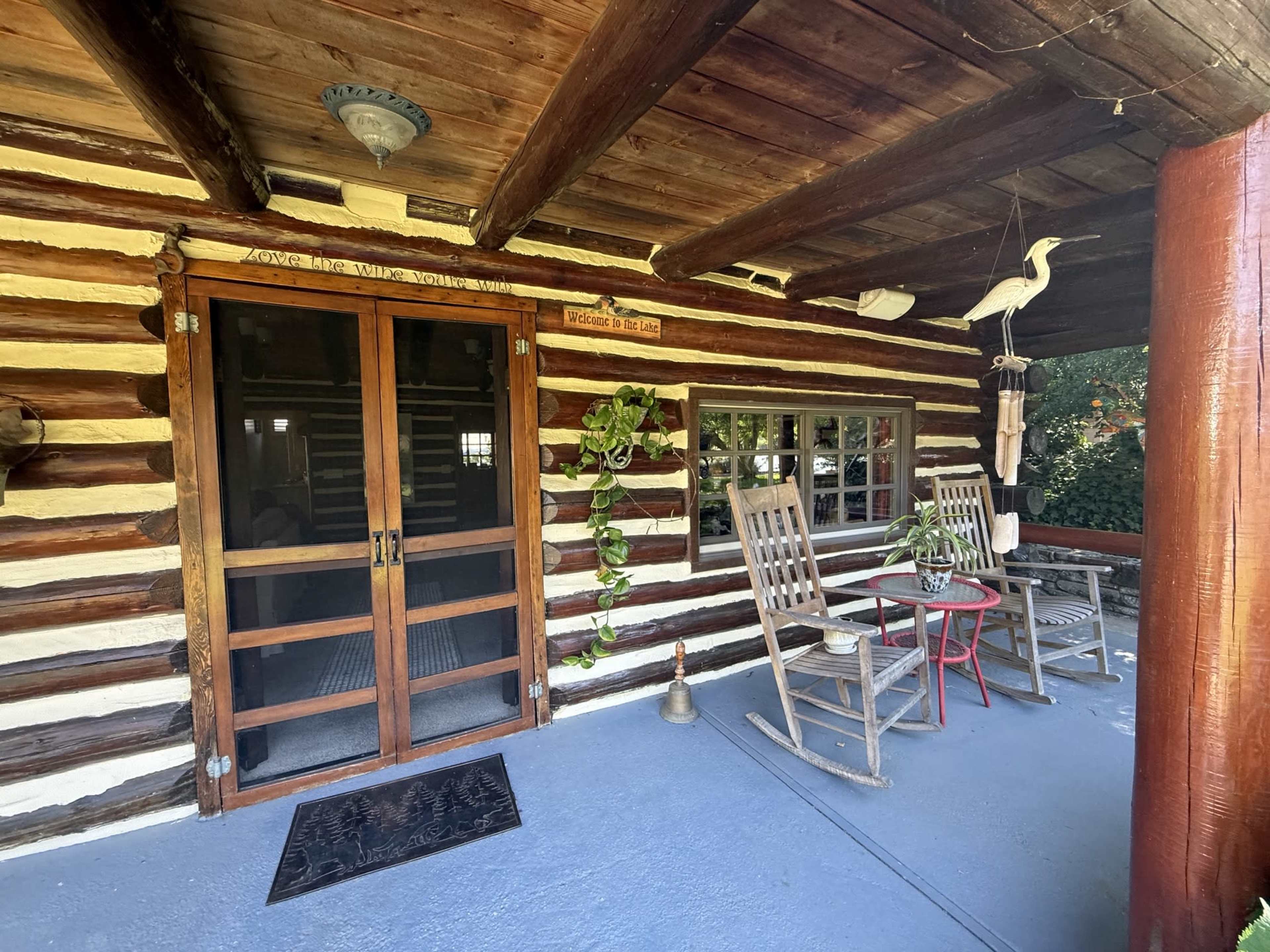 The image shows a rustic porch of a log cabin featuring wooden rocking chairs, a small table, and potted plants near a double door entrance.