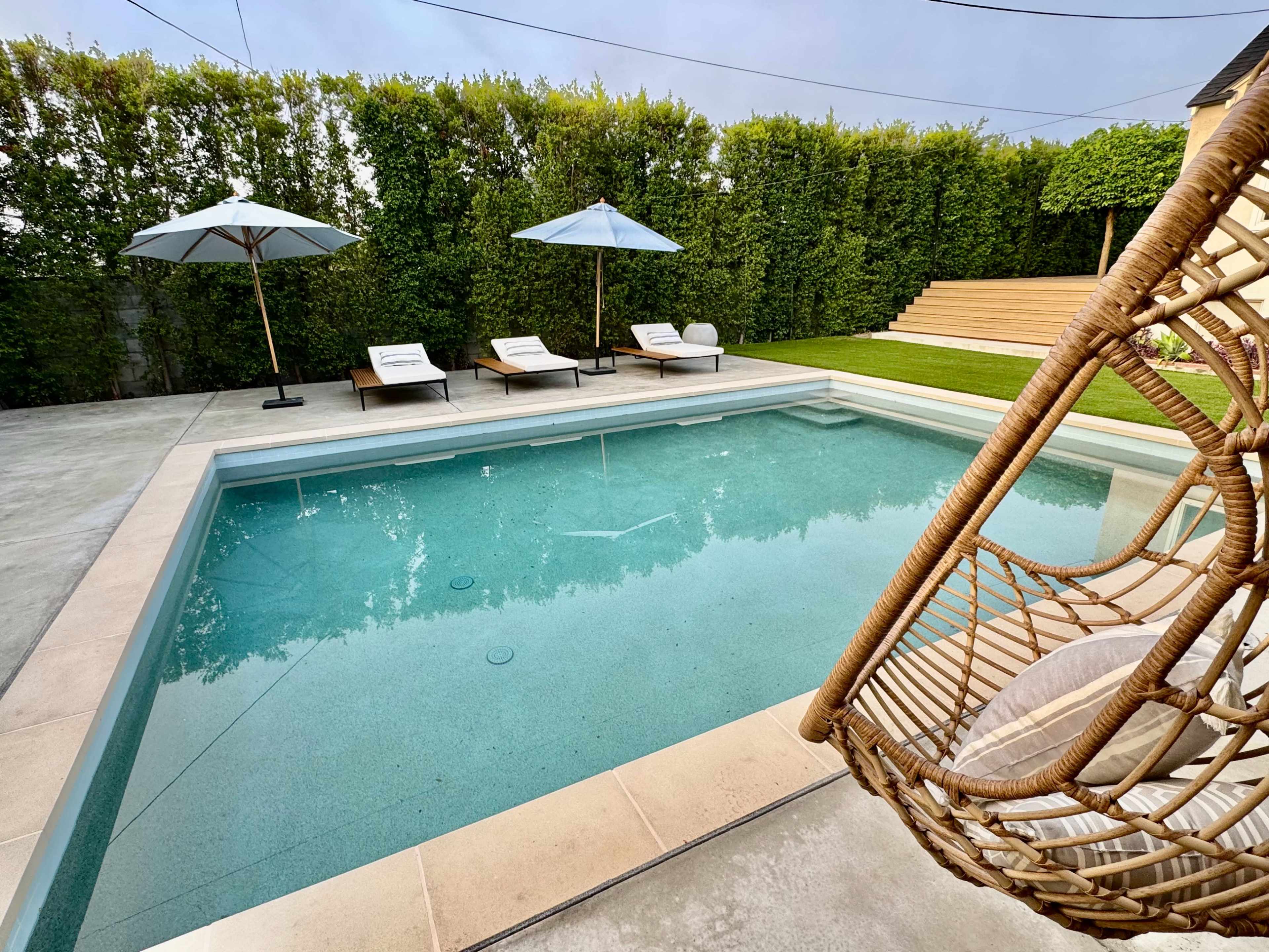 A rectangular swimming pool with a clear water surface is surrounded by lounge chairs and umbrellas, set against a backdrop of neatly trimmed hedges and grass.