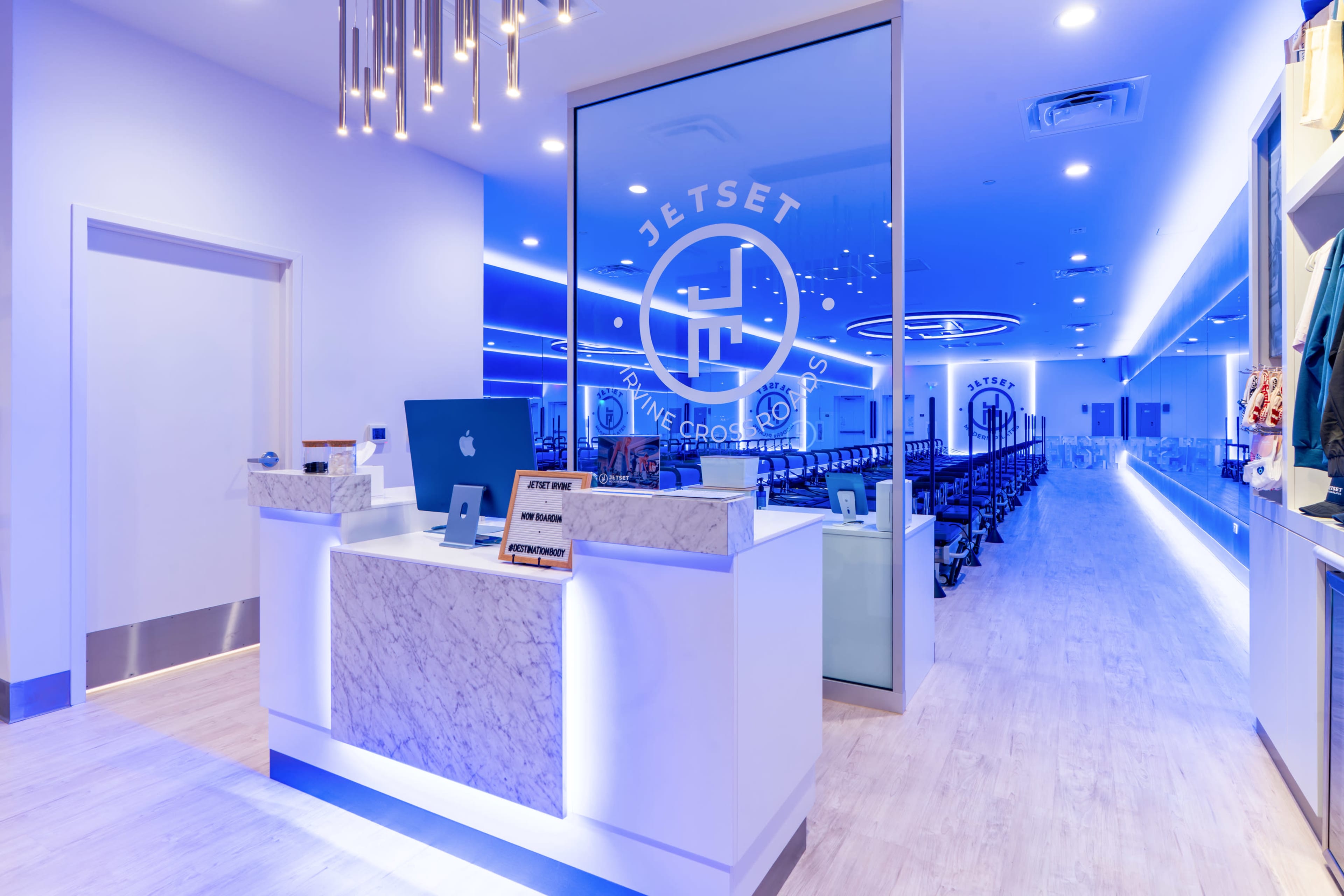 The image shows a modern reception area of a fitness facility with a sleek front desk, bright blue lighting, and a spacious workout area visible in the background.