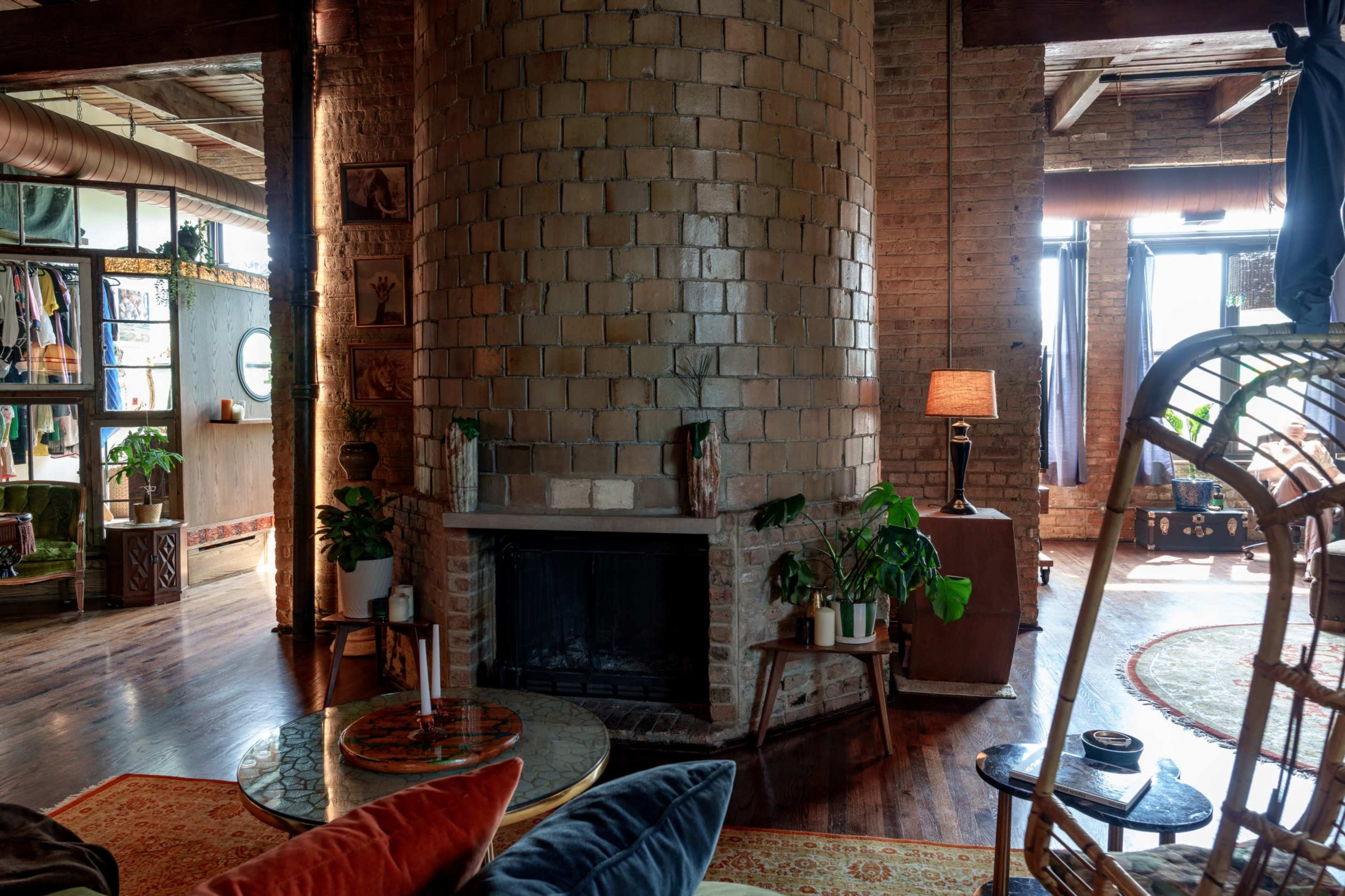 The image shows a cozy, industrial-style living space featuring a round brick column with a fireplace, large windows, and an eclectic mix of furniture and plants.