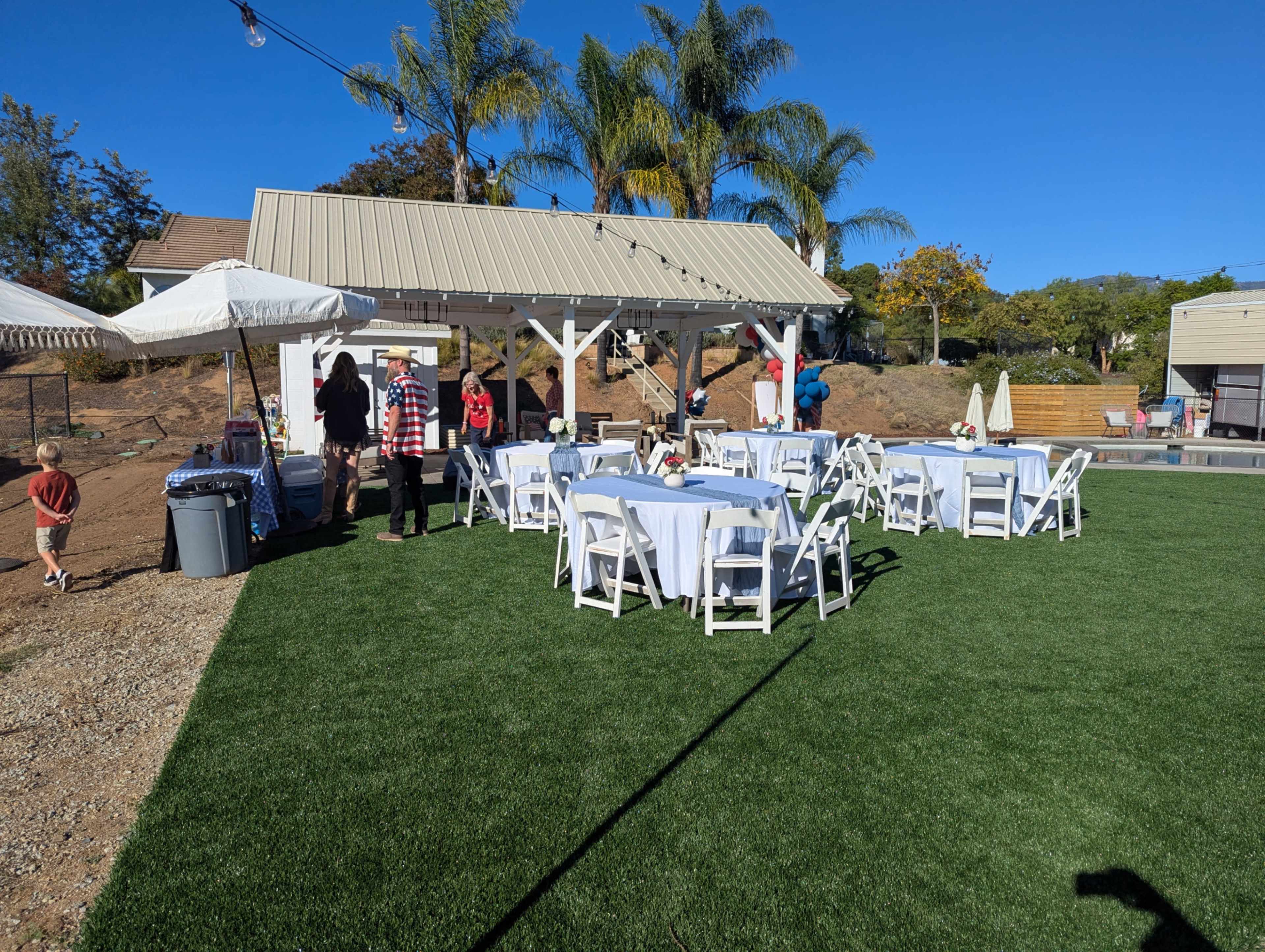 A patio area features several white tables and chairs set up on green artificial grass, with guests mingling near a covered serving area.