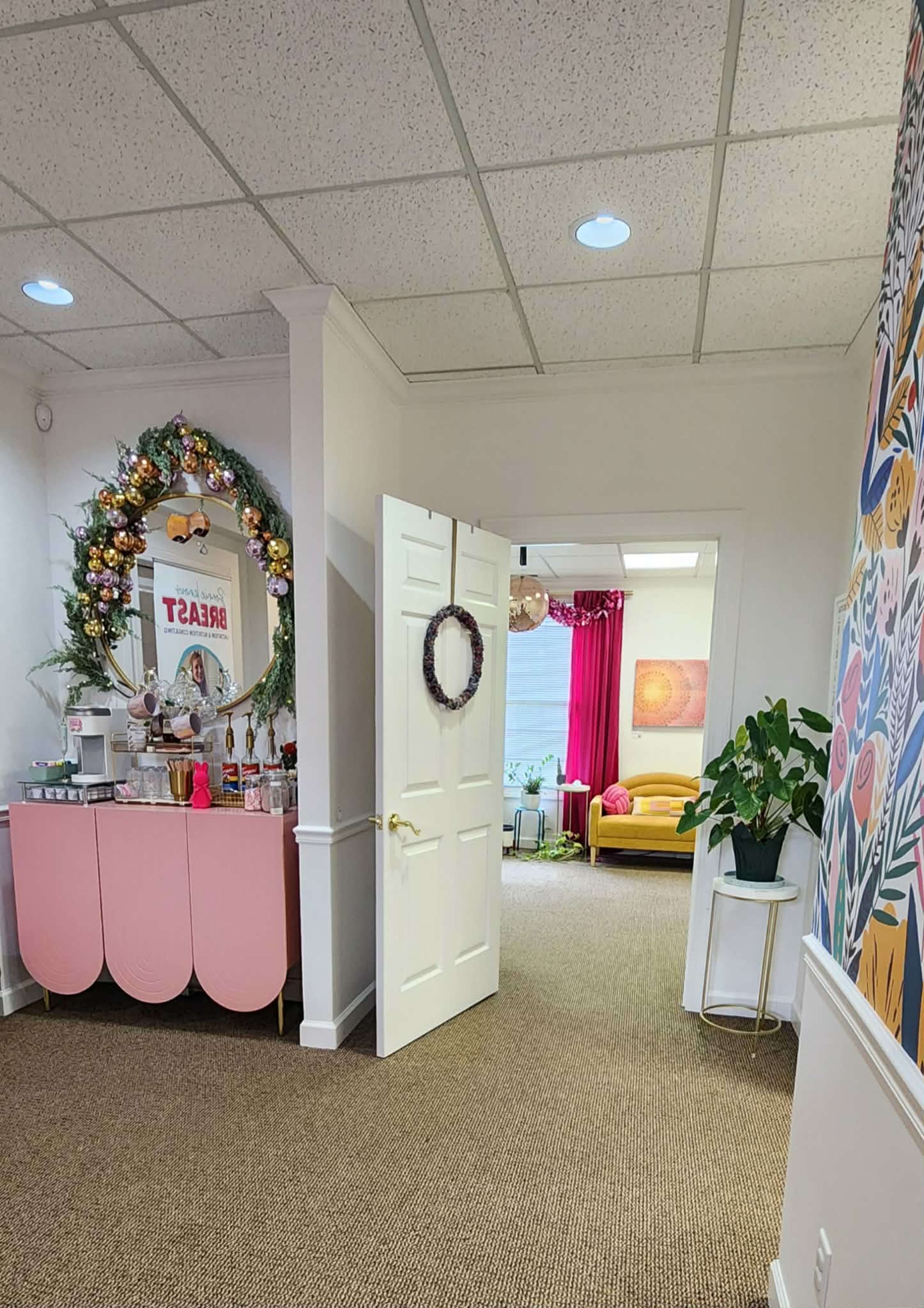 The image shows a hallway leading to a brightly colored room with a pink-curtained window and a yellow couch, while a pink side table decorated with a wreath and holiday decor is positioned nearby.