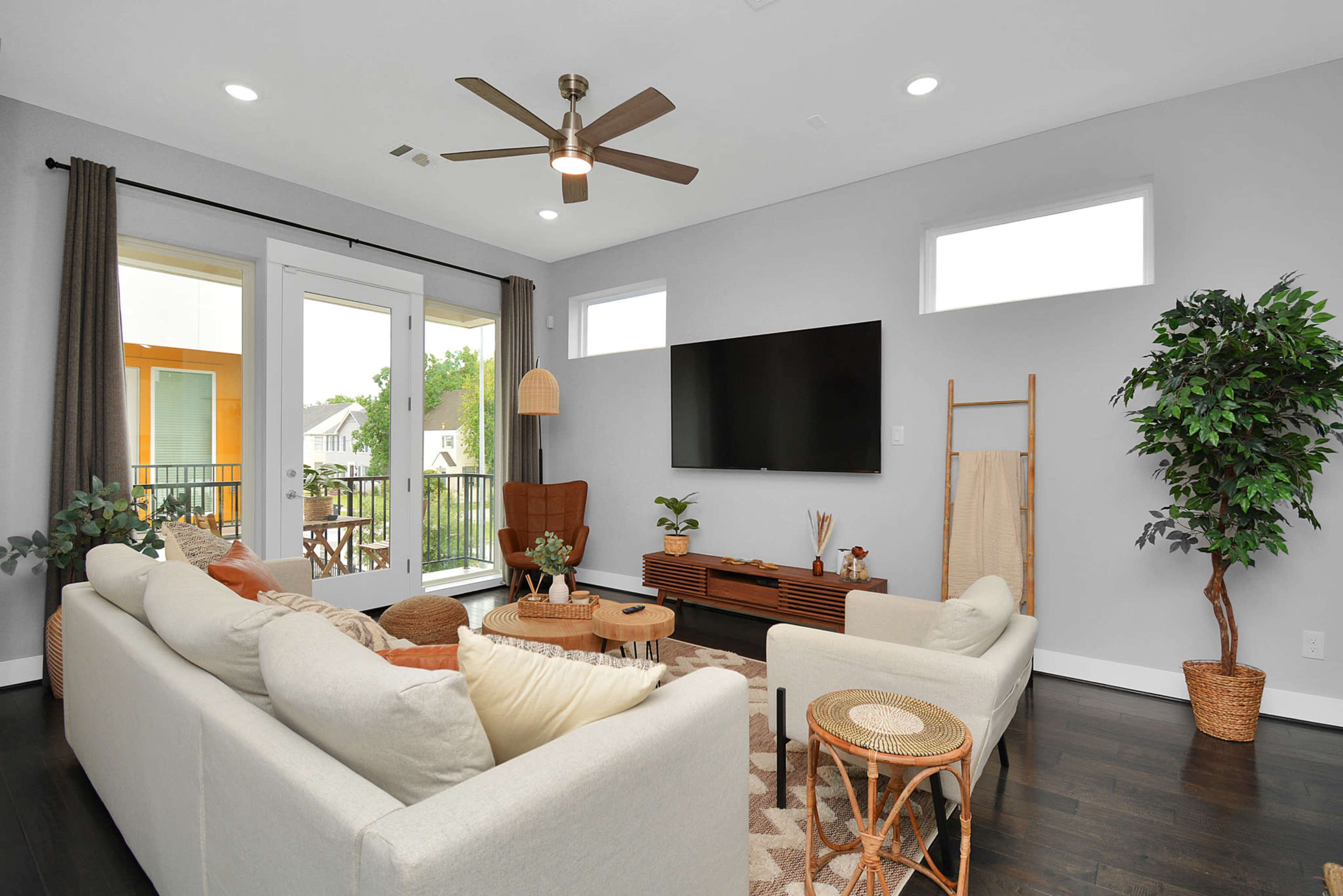 A modern living room features a large sofa, a flat-screen TV on a wooden console, a fan overhead, and large windows that open to a balcony.