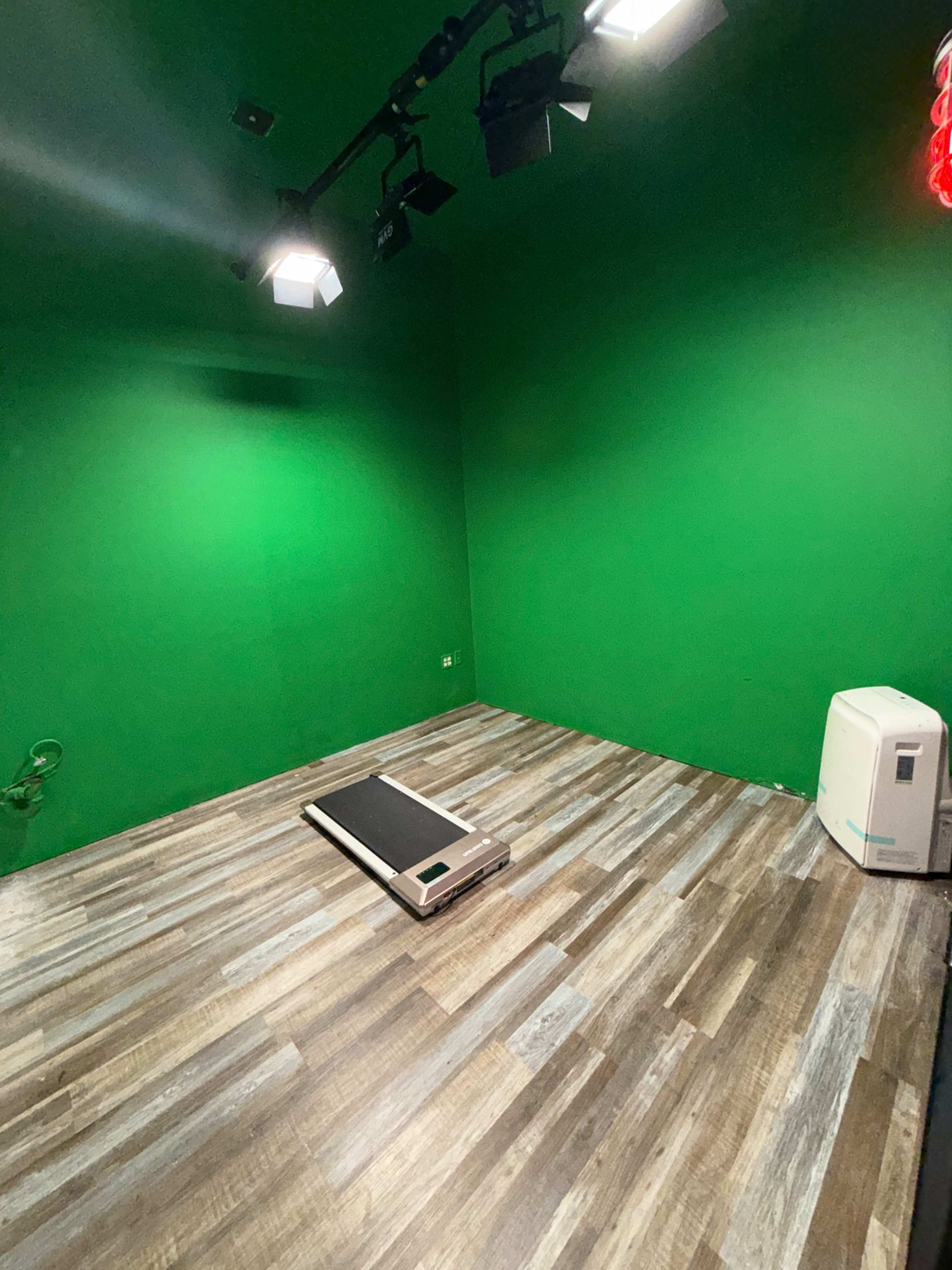 The image shows an empty room with green walls, a wooden floor, a treadmill in the center, and a white air conditioning unit in the corner.
