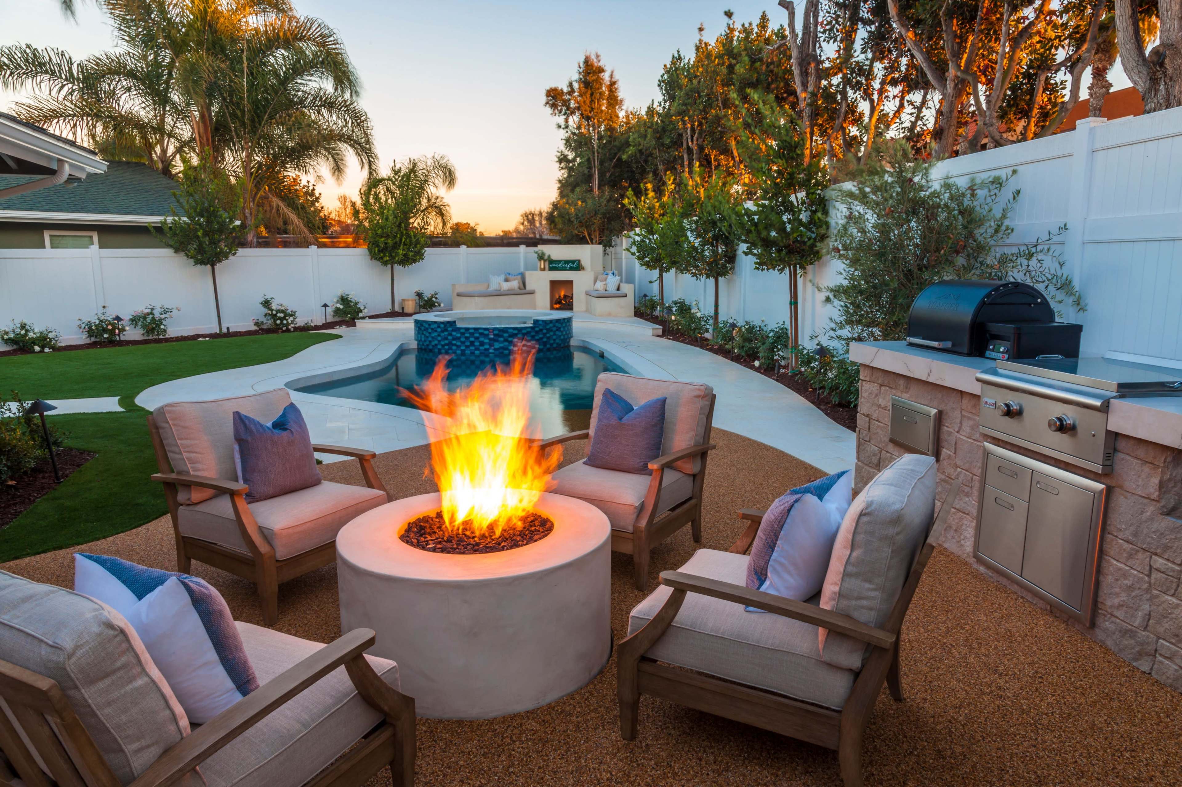 An outdoor patio with a fire pit surrounded by four chairs, a swimming pool in the background, and a barbecue grill nearby.