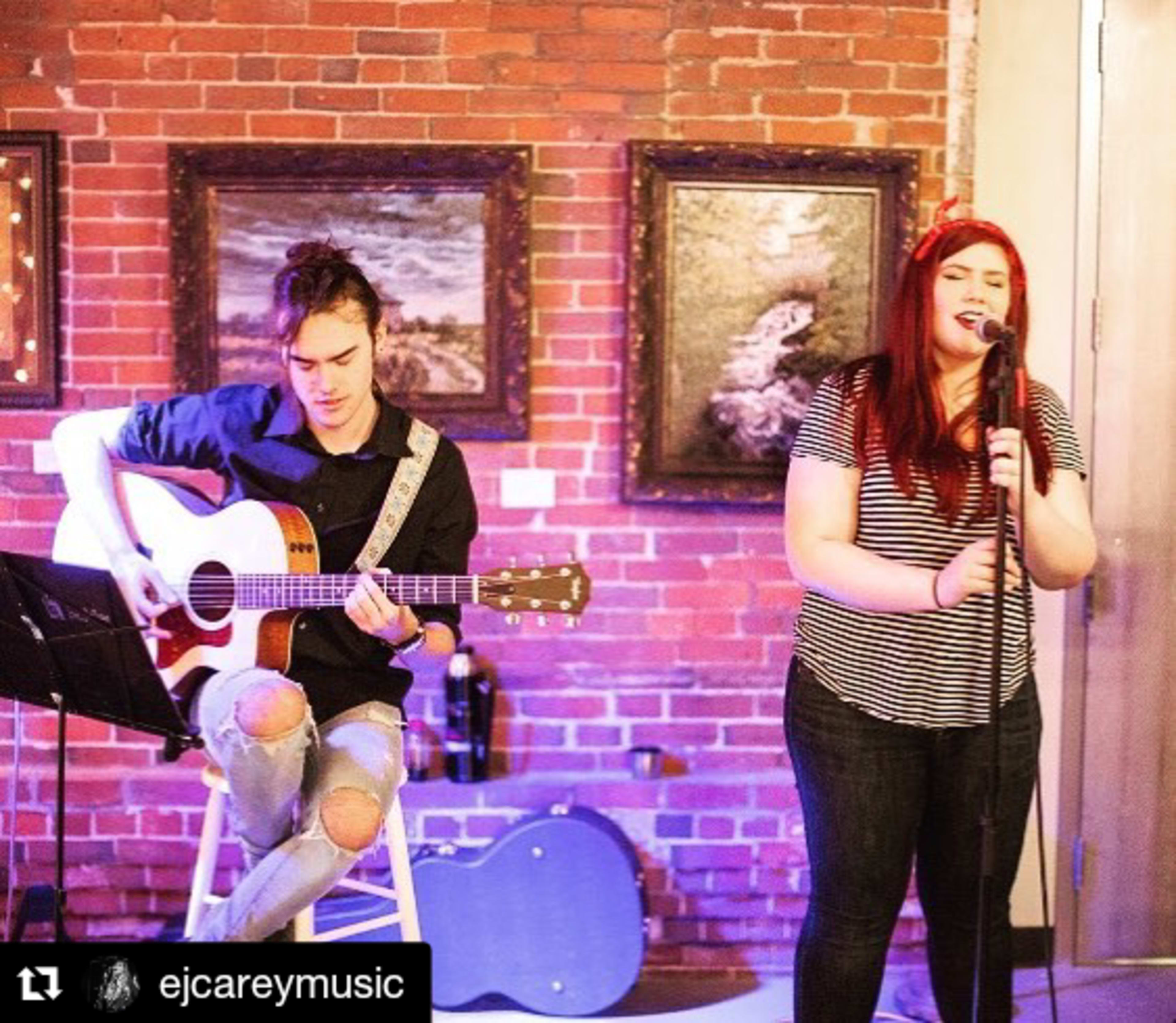 A man plays acoustic guitar while a woman sings into a microphone, with a brick wall and framed pictures in the background.
