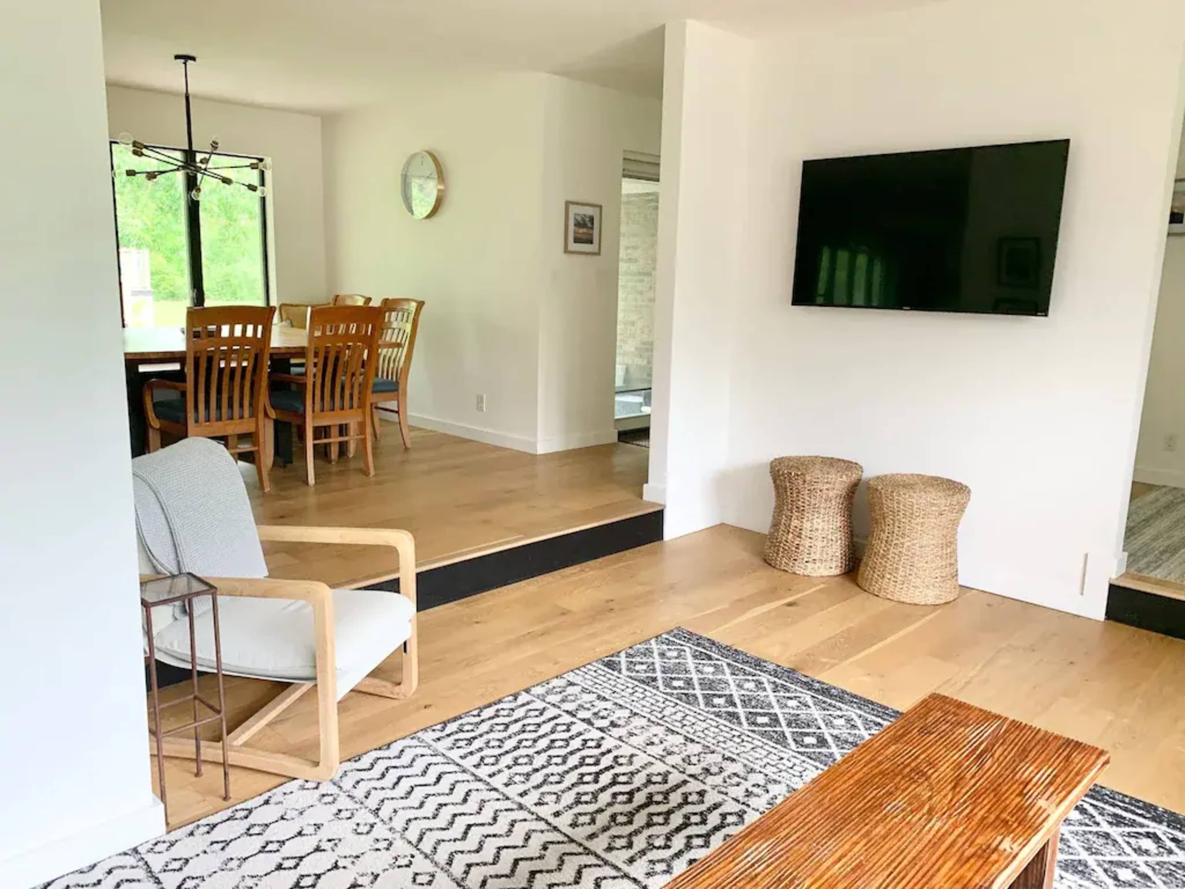 A light-filled living area featuring a chair, a wooden coffee table, a patterned rug, and a wall-mounted television, with a dining space visible in the background.