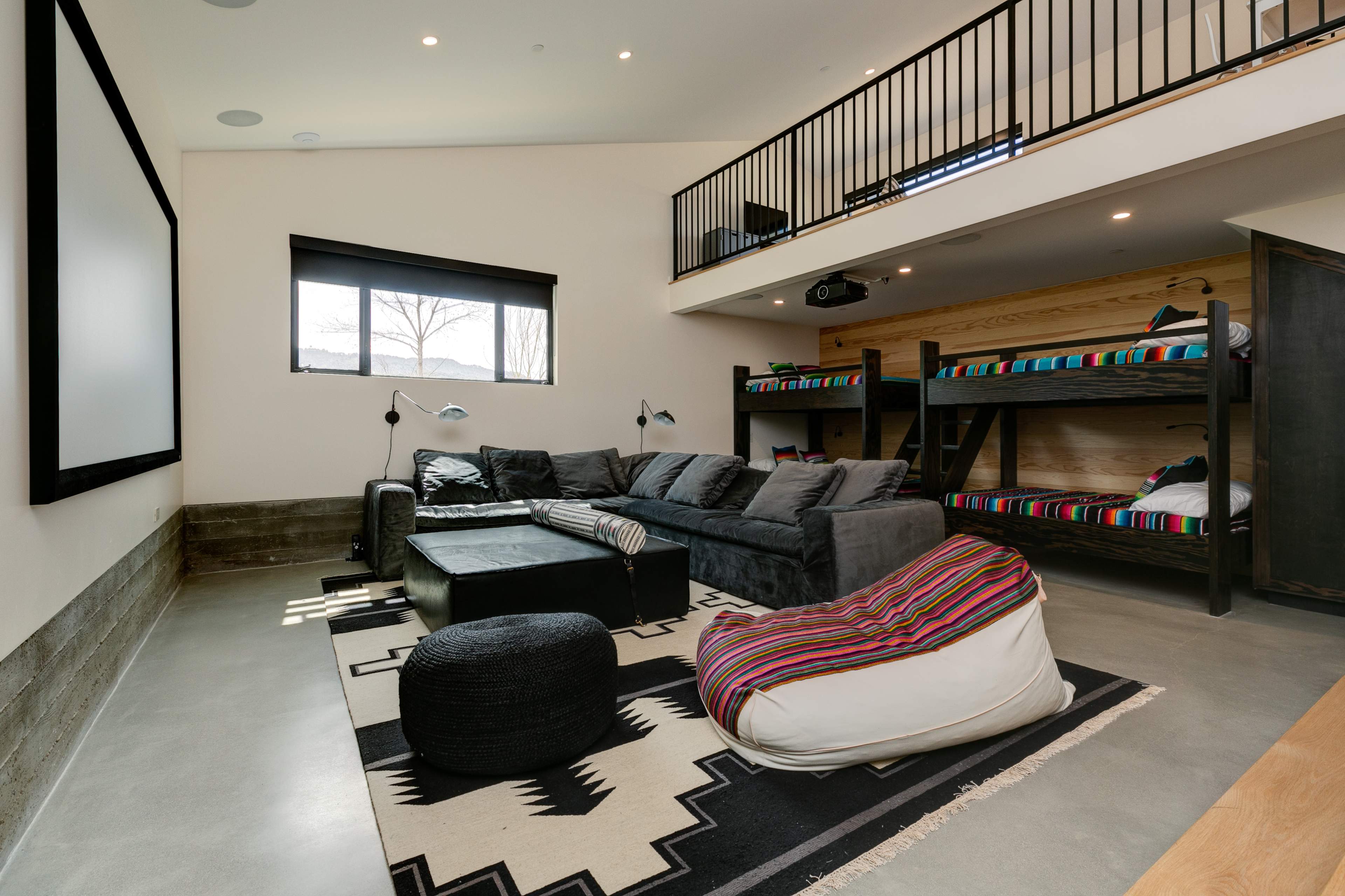 The image shows a modern, well-lit living area featuring a gray sectional sofa, a large screen, and a loft with bunk beds, all set on a concrete floor with a patterned rug.
