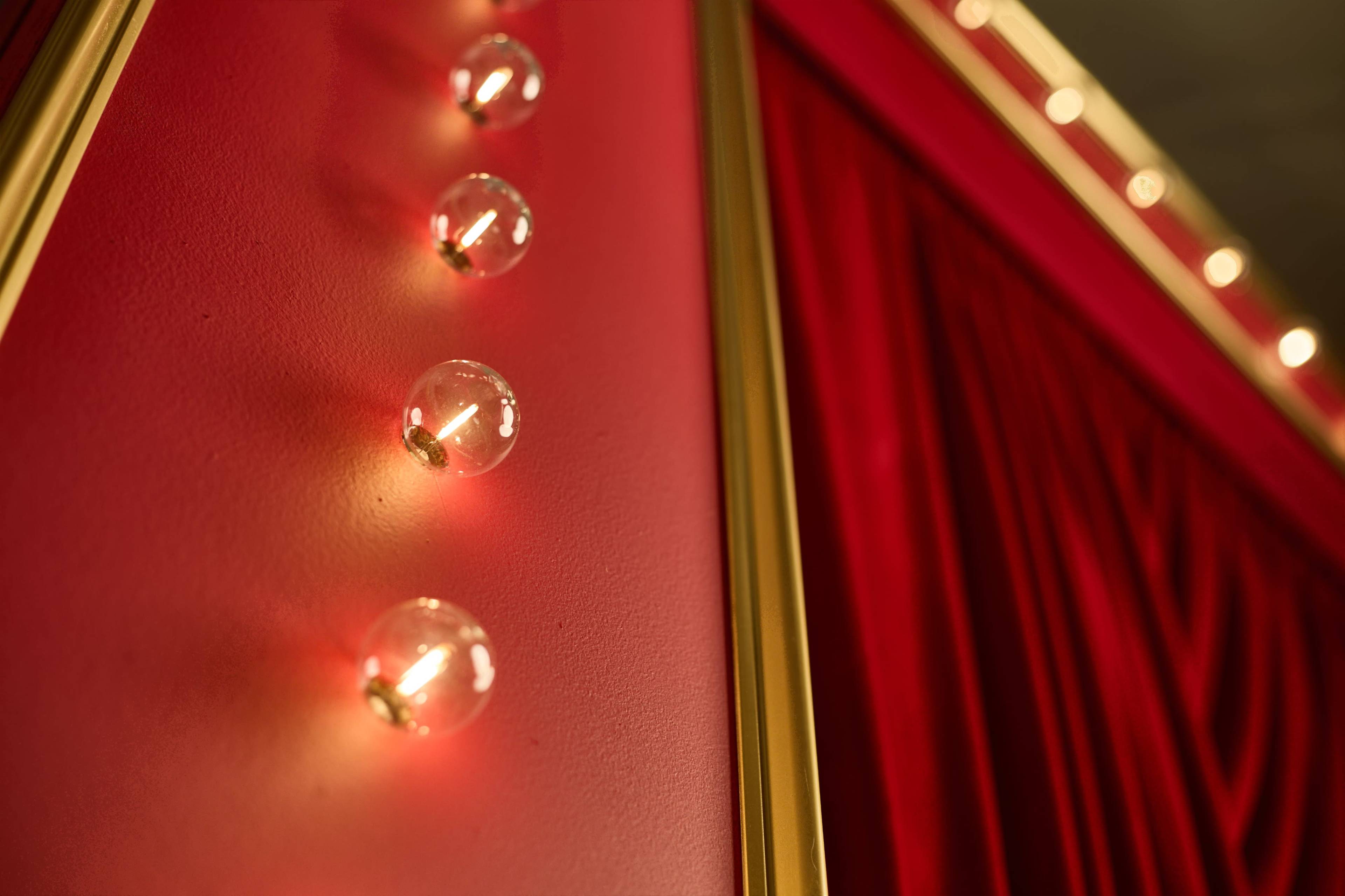 The image shows a close-up of illuminated round bulbs attached to a red wall with velvet drapery.