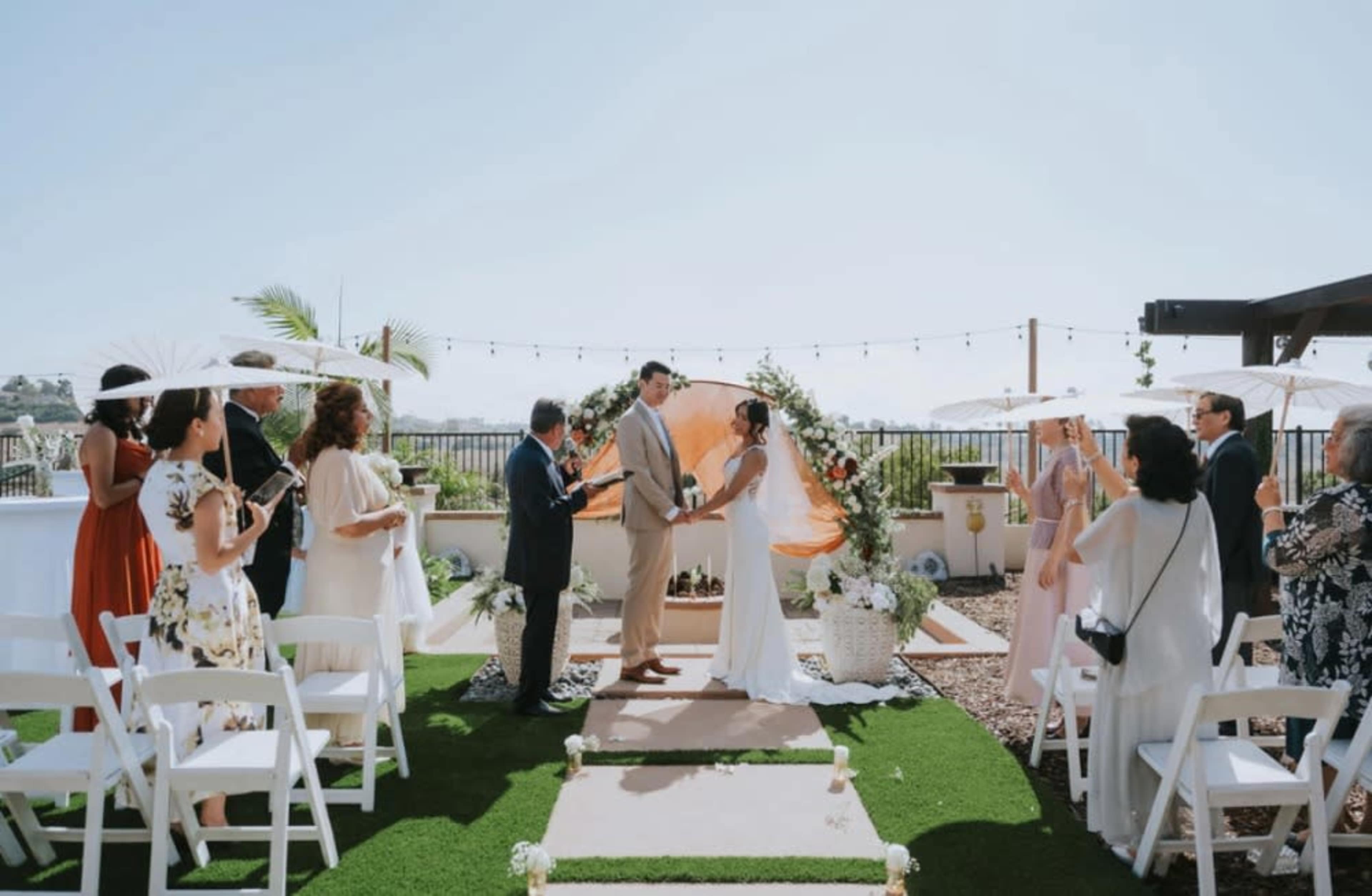 A couple exchanges vows under a floral arch on a patio, surrounded by seated guests with umbrellas and greenery.